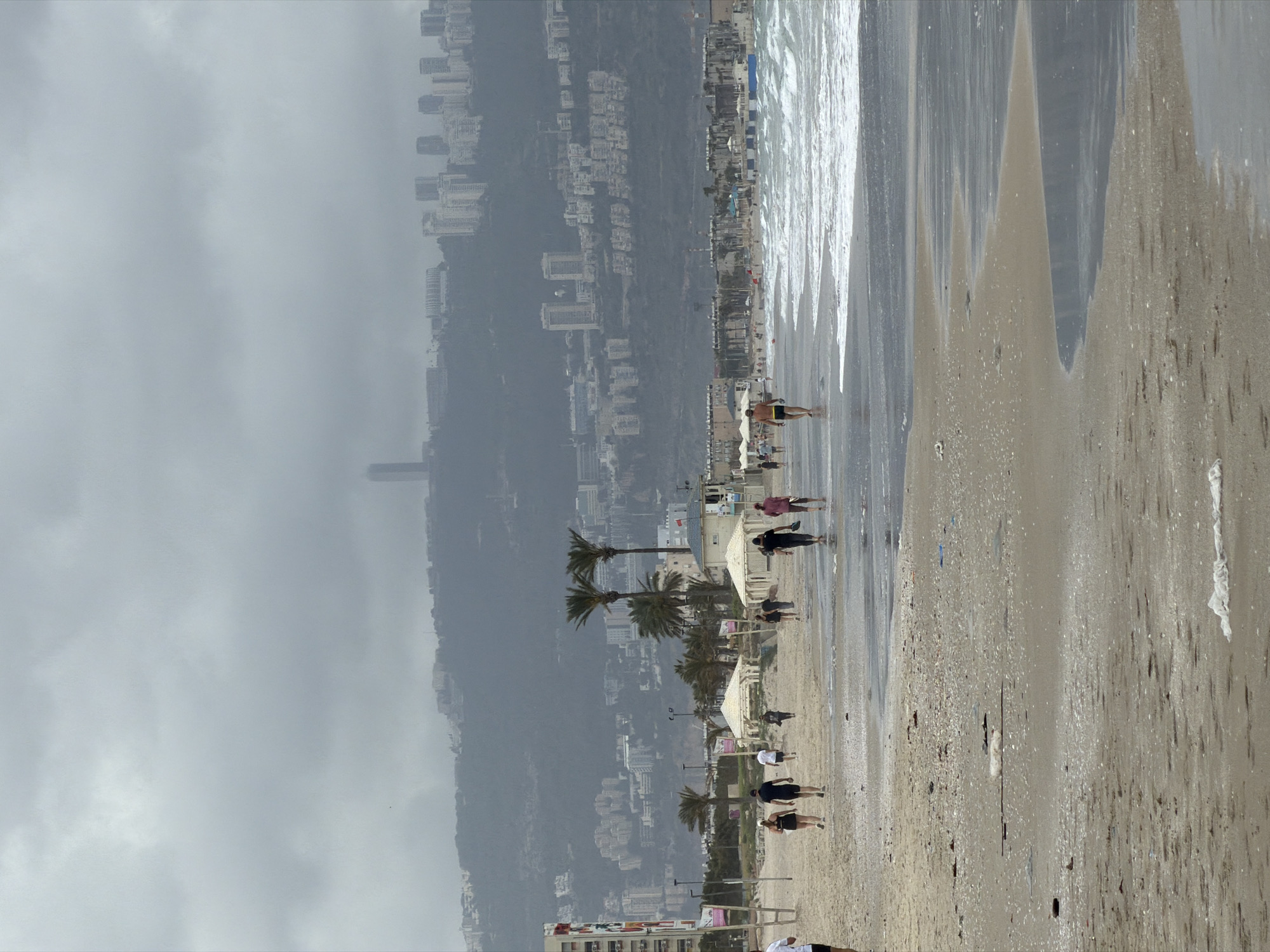 The image shows a beach scene in Kiryat Yam, Israel, under a cloudy sky. The coastline curves with the sea on the right. Waves are breaking along the shore. The beach is populated with people walking along the wet sand near the water's edge. Palm trees are sparsely distributed along the beach, indicating a landscaped area. Behind the beach, the city rises up a hill or slope. Buildings of varying sizes and styles are densely packed. The cloudy weather obscures the upper reaches of the cityscape.
