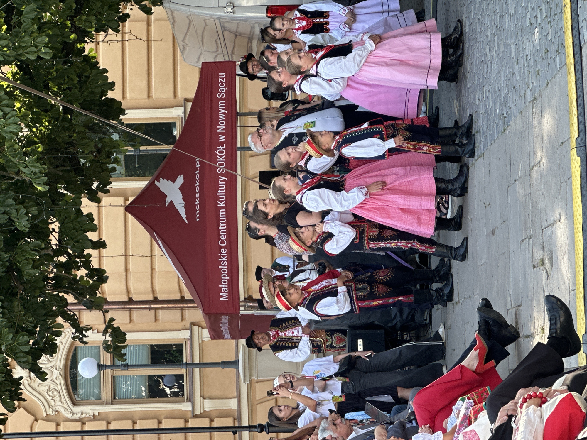 A group of children and adults are participating in a cultural event, possibly a festival or performance, in front of a building in the city of Nowy Sącz, Poland. Many of the participants, particularly the children, are dressed in traditional folk costumes. These costumes include embroidered vests, white shirts, and dark pants or skirts, with some boys wearing straw hats. Several of the children are wearing bright pink skirts. Adults in the background are also dressed in similar traditional attire or more casual clothing. A red tent with text indicating "Małopolskie Centrum Kultury SOKÓŁ w Nowym Sączu" (Malopolska Cultural Center SOKOL in Nowy Sacz) is set up behind the performers. The event is taking place outdoors on a paved area, with trees and a building visible in the background. Some spectators are seated in the foreground, observing the event, while others are standing and taking photos. The overall scene suggests a lively cultural gathering in a public square in Nowy Sącz.