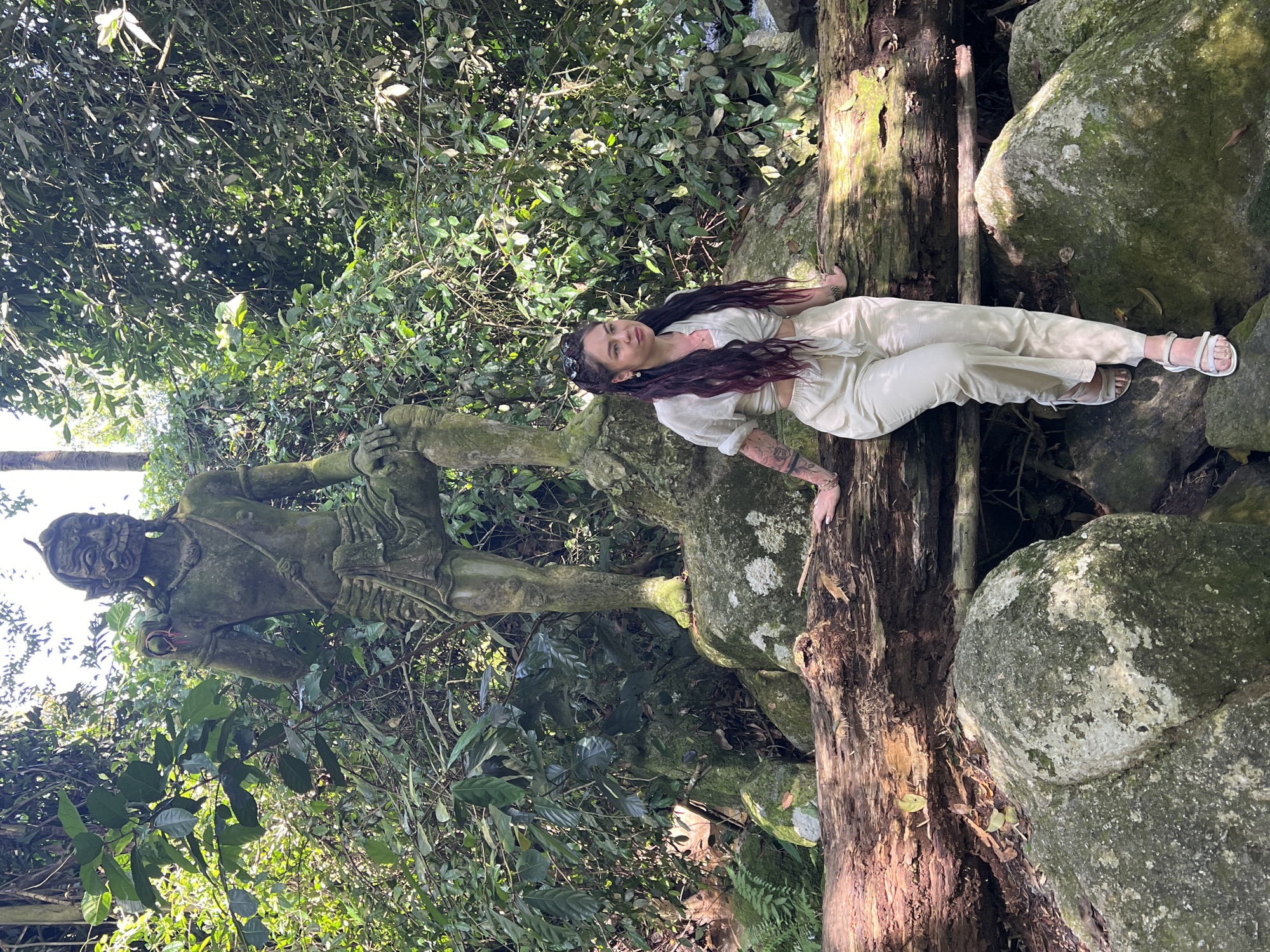 A woman in a light-colored outfit with a tied-front top and loose pants is sitting on a fallen log in front of a moss-covered stone statue of a warrior. The statue is in a jungle environment with lush green foliage and dappled sunlight. Large rocks are scattered around the base of the statue and the log. The woman has long, dark hair with reddish highlights and is wearing sandals. Her left arm rests on the log, and her right hand is also near the log. The statue appears to be ancient and weathered. The scene is set in the city of Ban Bang Khao, Thailand.