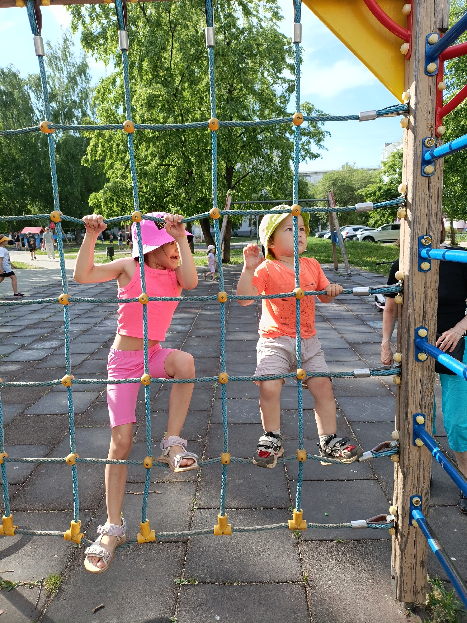 Two young children are playing on a rope climbing structure in a playground. The girl, on the left, is wearing a pink tank top, pink shorts, and pink hat. She is climbing the net with one leg up. The boy, on the right, is wearing an orange shirt, tan shorts, and a green hat. He is holding onto the rope. An adult is standing next to the climbing structure. The playground has a gray rubber surface and is located in a park with trees and other playground equipment. The location is the city of Chaikovsky, Russia.