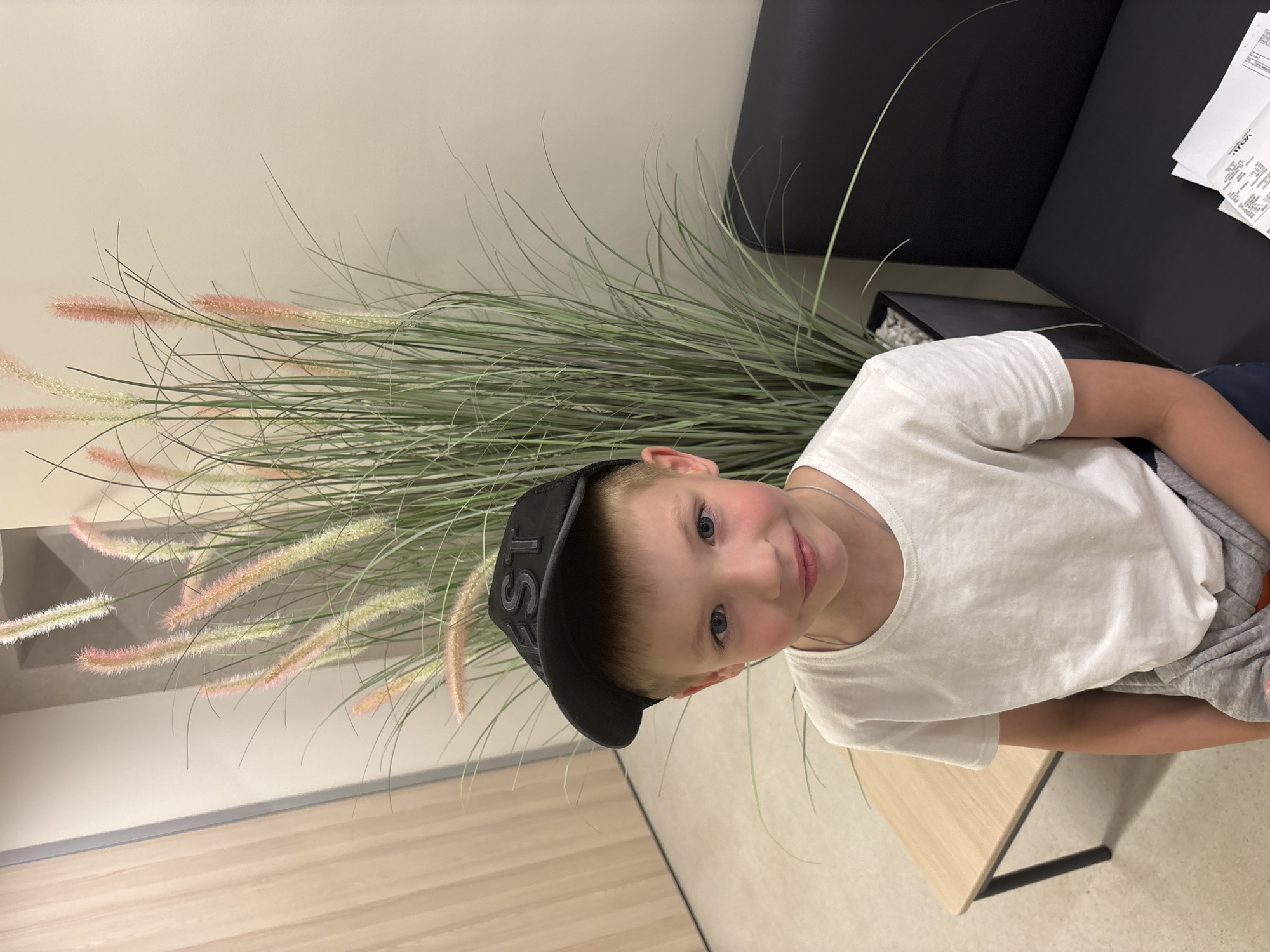 A young boy wearing a black baseball cap with the letters "ST" on it and a white t-shirt is seated indoors. He has light brown hair and blue eyes, and is looking towards the camera with a slight smile. Behind him, a tall artificial plant with feathery plumes is visible. To his left is a small wooden table with black metal legs. To his right is a black bench or chair, and a stack of papers is on the floor next to it. The environment appears to be a waiting room or a similar indoor space with neutral-colored walls and flooring. There is no identifiable information about the location in the city of Izhevsk, Russia.