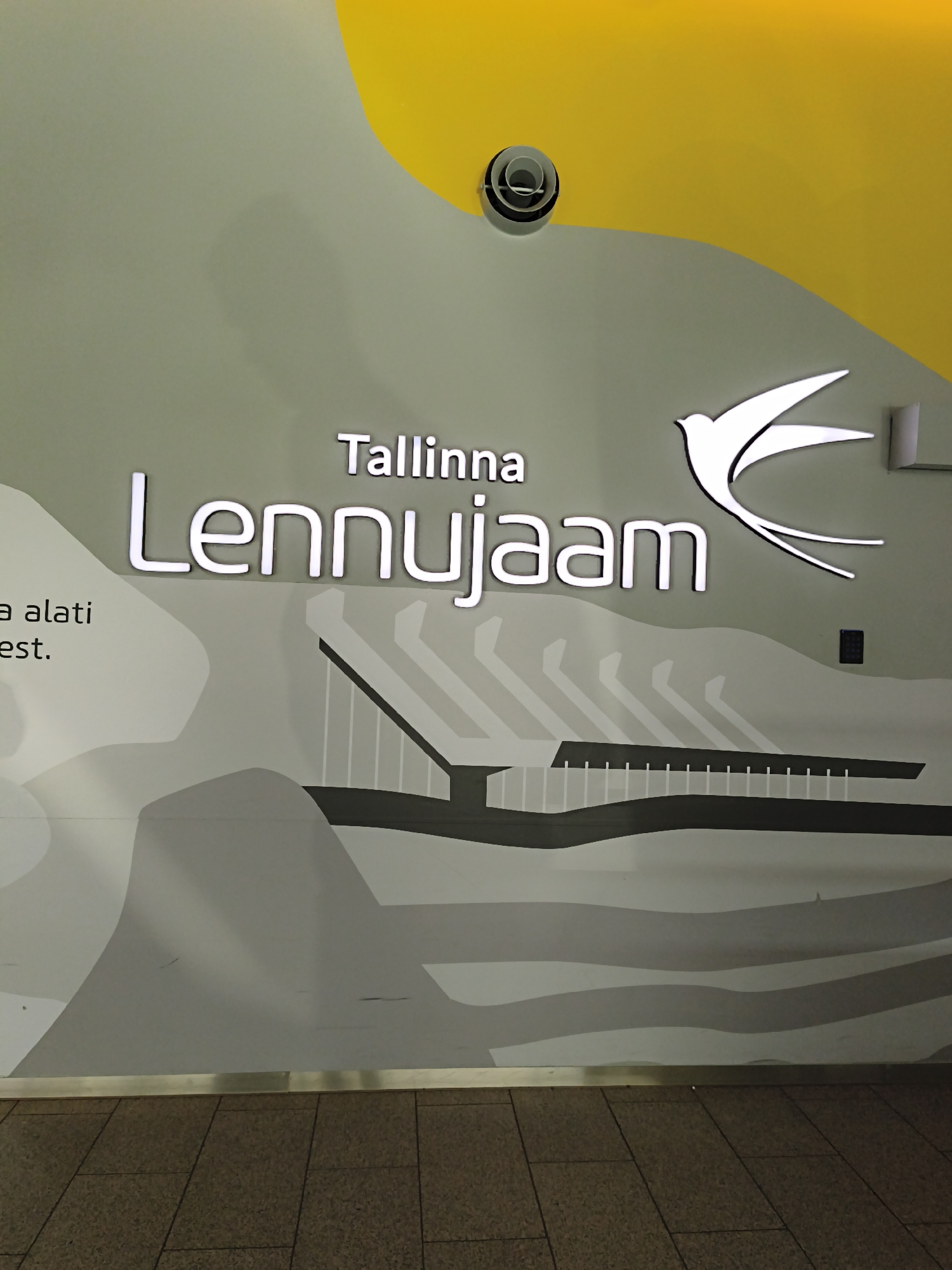 The image shows a wall mural at Tallinn Airport (Lennujaam) in Tallinn, Estonia. The mural features the words "Tallinna Lennujaam" in large, illuminated lettering, accompanied by a stylized white bird in flight. The background of the mural is a gradient of light gray and green, with abstract wavy shapes and a depiction of a bridge structure. A yellow curved shape is visible at the top of the wall, and a circular ventilation fixture is mounted on the wall near the yellow area. A small dark square device, possibly an intercom or access panel, is also present on the wall. The bottom of the image shows a tiled floor in a muted brown color, separated from the wall by a metallic trim. The overall scene is an interior shot of the airport terminal, highlighting its branding and design.