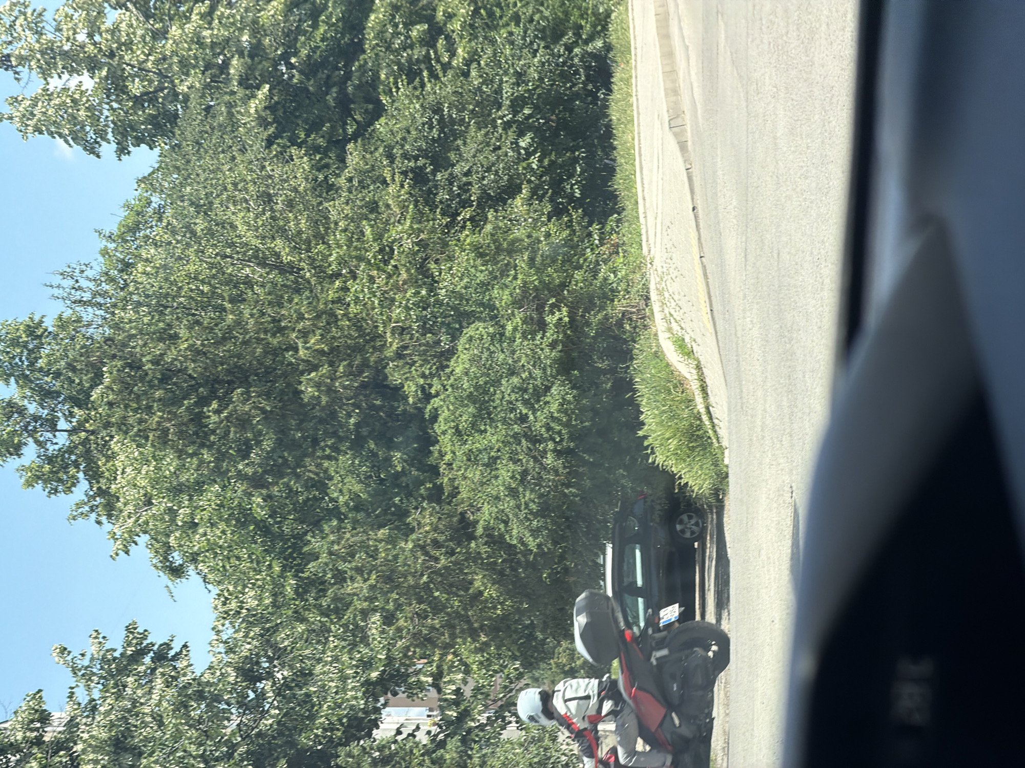 In Sofia, Bulgaria, on a bright, sunny day, a person wearing a light blue helmet and a white and grey motorcycle jacket rides a red motorcycle with a black storage box. The motorcycle is moving on a paved road. To the right of the road, there is a black car parked. The background is filled with lush green trees and vegetation. In the foreground, a portion of a car's dashboard is visible, showing a digital display. The overall scene depicts an urban outdoor environment with a focus on a motorcycle and a parked car against a backdrop of natural greenery.