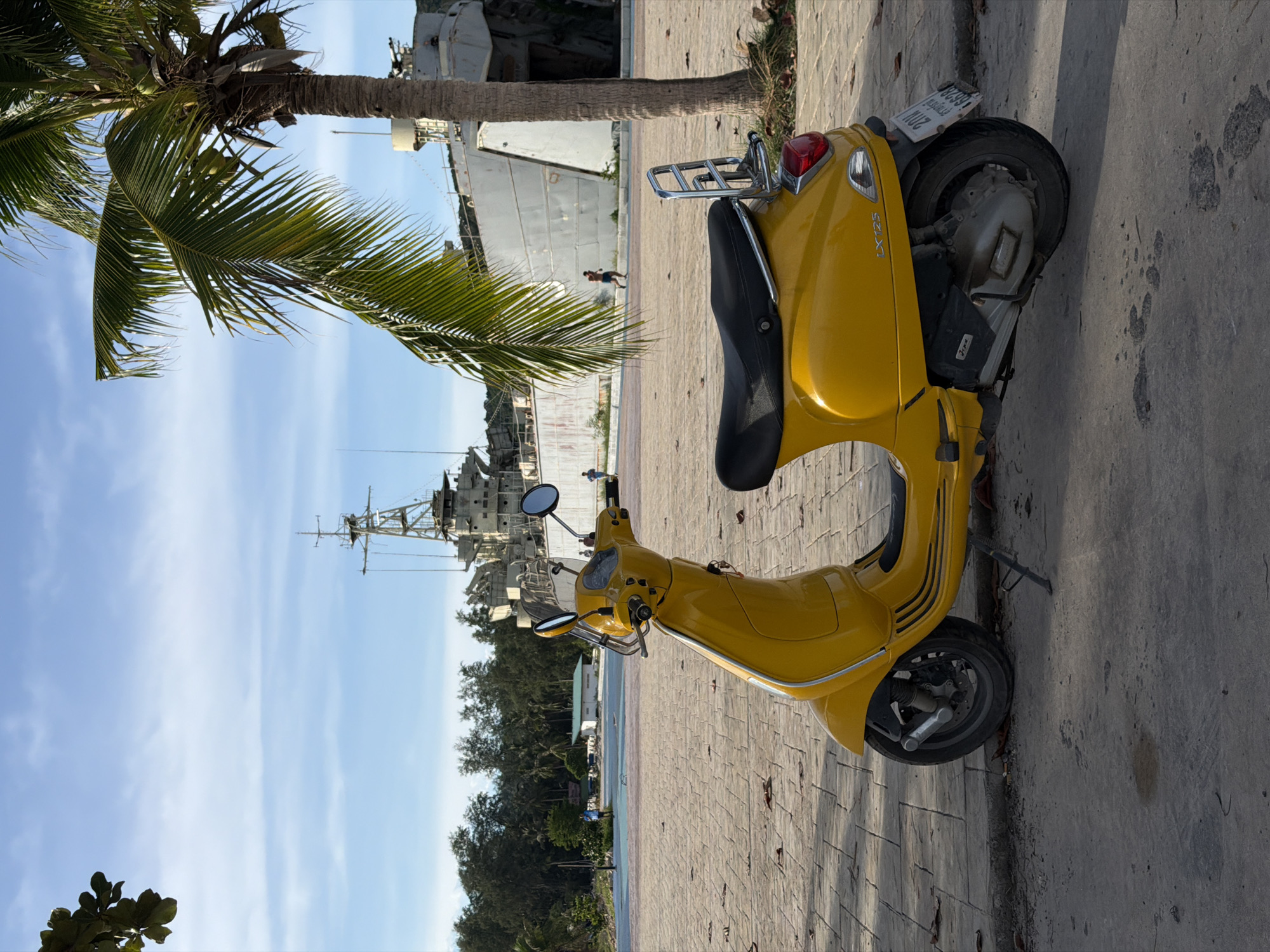 A bright yellow Vespa scooter is parked on a paved walkway in front of a large, grey ship that appears to be permanently docked. The ship is old and weathered, with visible signs of disrepair. Behind the ship, lush green trees and a palm tree frame a bright blue sky with scattered white clouds. Several people are visible in the background, walking near the ship and a small, light-colored building with a green roof. The ground in the foreground is a mixture of concrete and paving stones, with fallen leaves scattered across it. The scene suggests a tropical or subtropical location, likely in the city of Ko Pha Ngan, Thailand, given the presence of the large ship which could be a museum or part of a historical site.