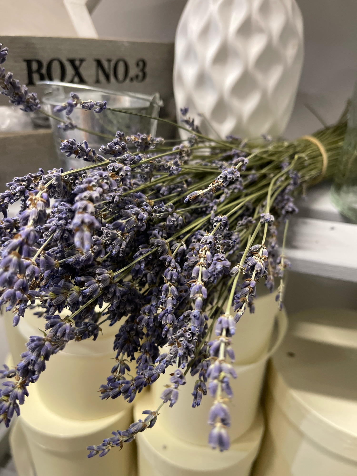 A close-up, shallow depth of field shot features a bunch of dried lavender stalks bound with twine. The lavender is predominantly purple and green, with some stalks appearing dried and slightly brittle. In the background, a white, diamond-patterned vase is visible, and to the left, a rustic wooden box with the text "BOX NO.3" printed on it. Below the lavender, several cream-colored cylindrical containers are stacked. The scene appears to be indoors, possibly in a shop or home, with shelving visible in the background. The lighting is soft and diffused, creating a slightly hazy atmosphere. The location is the city of Kommunarka, Russia.