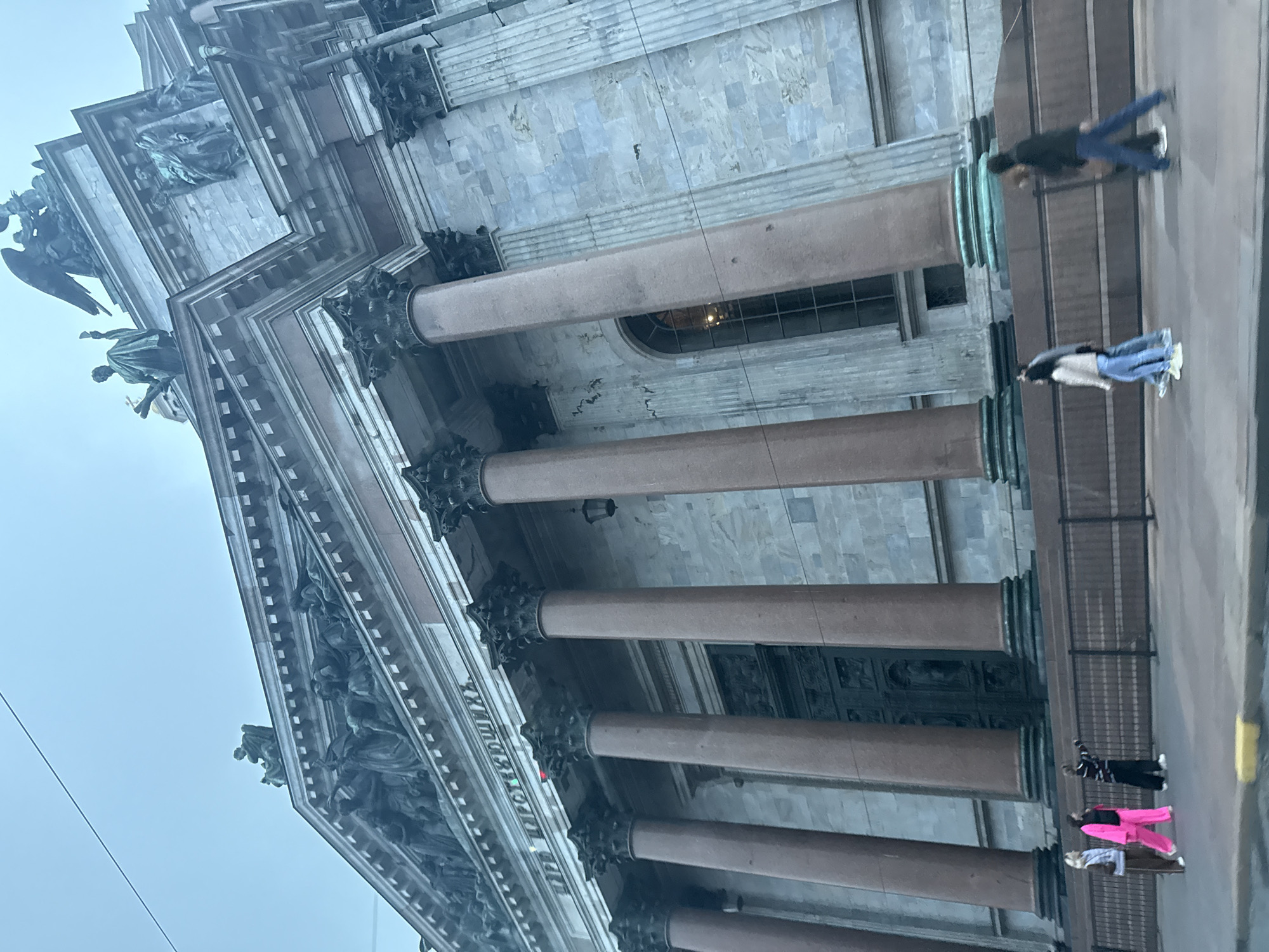 The image shows a low-angle, exterior view of St. Isaac's Cathedral in Saint Petersburg, Russia. The massive neoclassical building features a gray stone facade adorned with columns, sculptures, and architectural details. In the foreground, a group of individuals is walking along a paved pathway, separated from the cathedral by a dark metal fence. The sky is overcast, contributing to a muted and somewhat somber atmosphere. The architecture of St. Isaac's Cathedral is a prominent subject, with its imposing columns and decorative elements dominating the frame. The people are small figures in the composition, walking in various directions, indicating a public space. The overall scene is urban, with the grandeur of the cathedral contrasted by the ordinary activity of pedestrians.