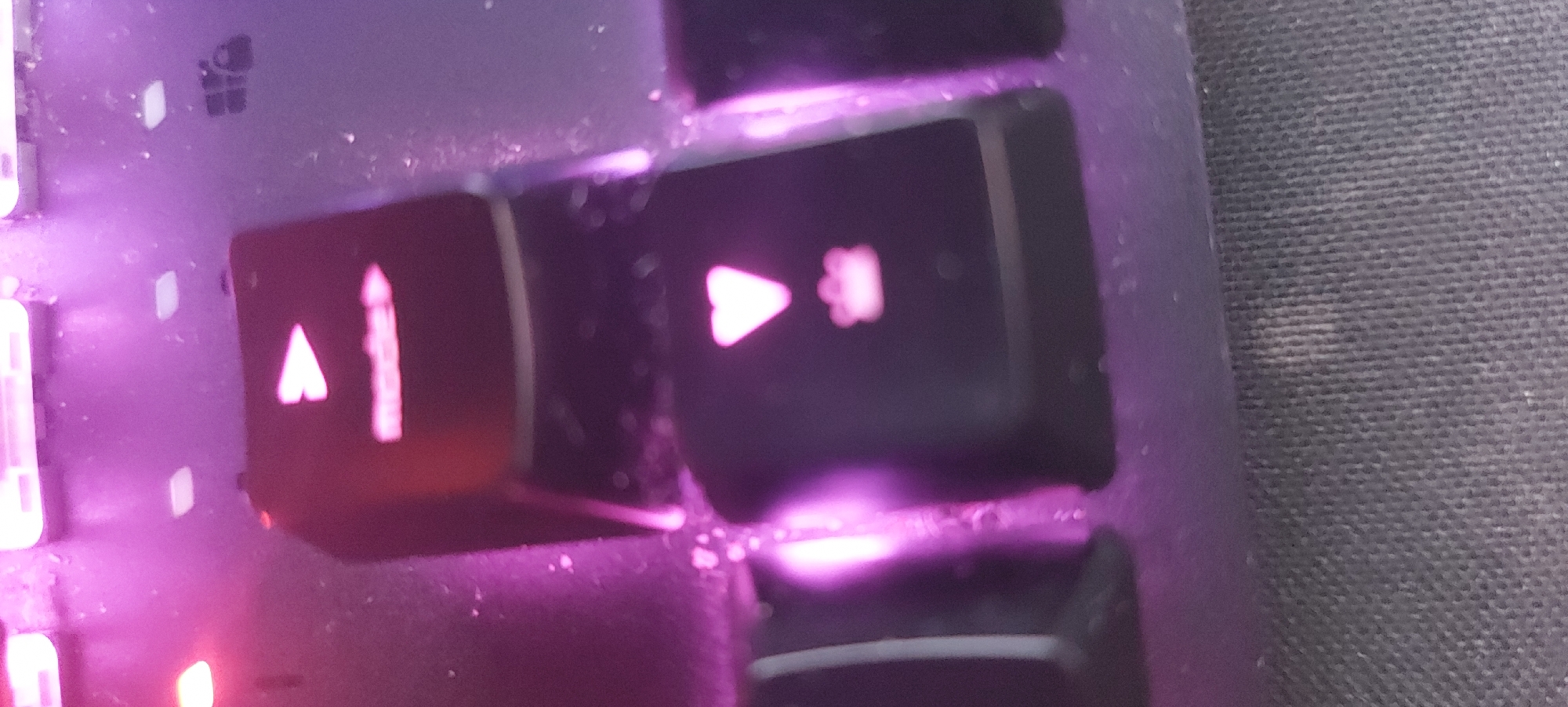 The image shows a close-up of a keyboard with illuminated keys. The keys are black, with illuminated symbols in pink. The symbols visible are an upward arrow with a vertical line next to it, and a right-pointing arrow with a symbol resembling a flower or asterisk next to it. The keyboard is dusty. A portion of a gray surface with a textured pattern is visible to the right of the keyboard. The location in Tashkent, Uzbekistan is not discernible from the image.