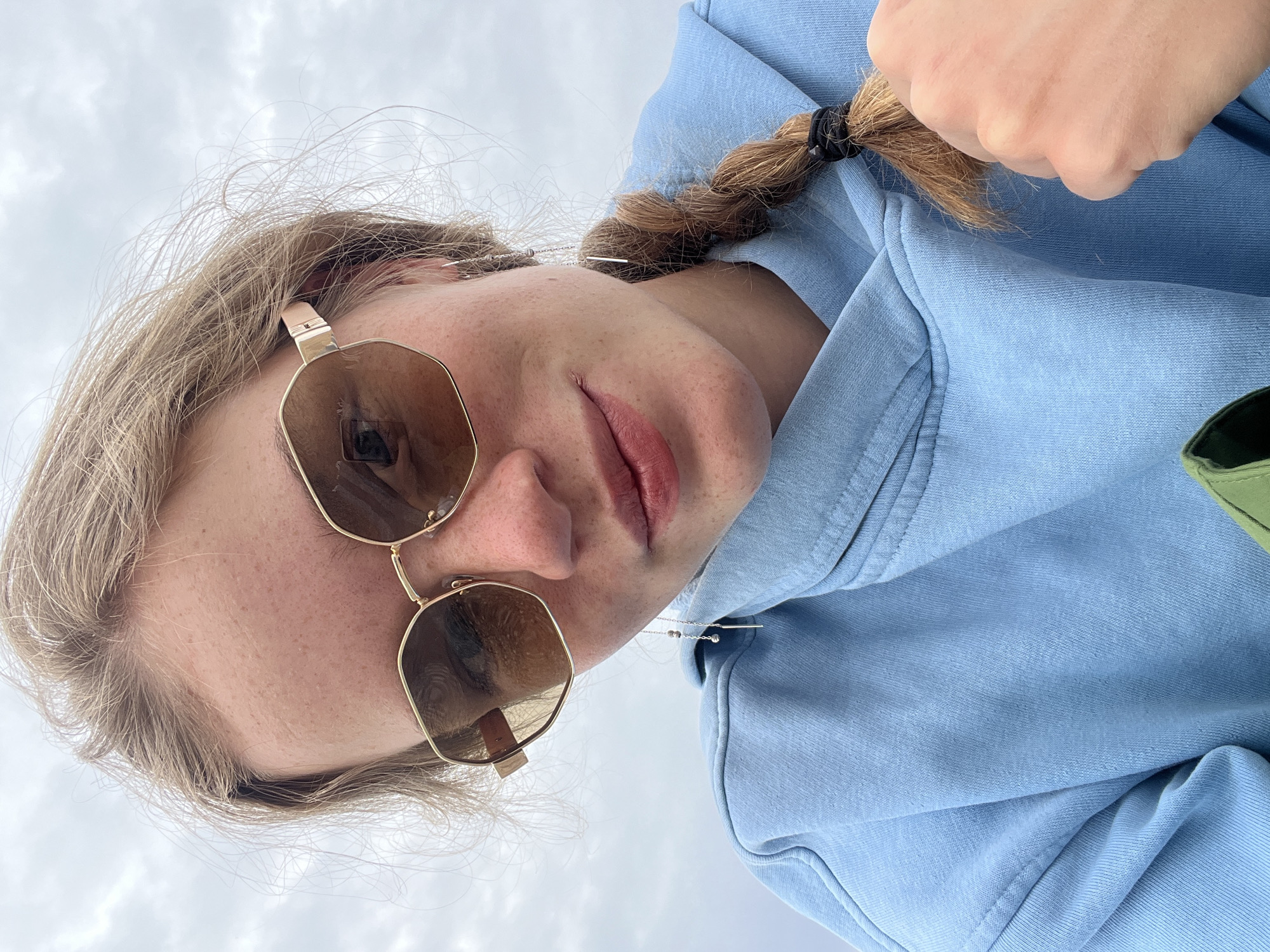 A selfie shows a woman with light blond hair and fair skin. She is wearing octagonal sunglasses with gold frames and brown lenses. Her hair is partially braided, and she's wearing a light blue hoodie. In the background is an overcast sky. The photograph's orientation suggests it was taken from a low angle, looking up at the subject. No information exists to suggest this image was taken in Boreti, Montenegro.
