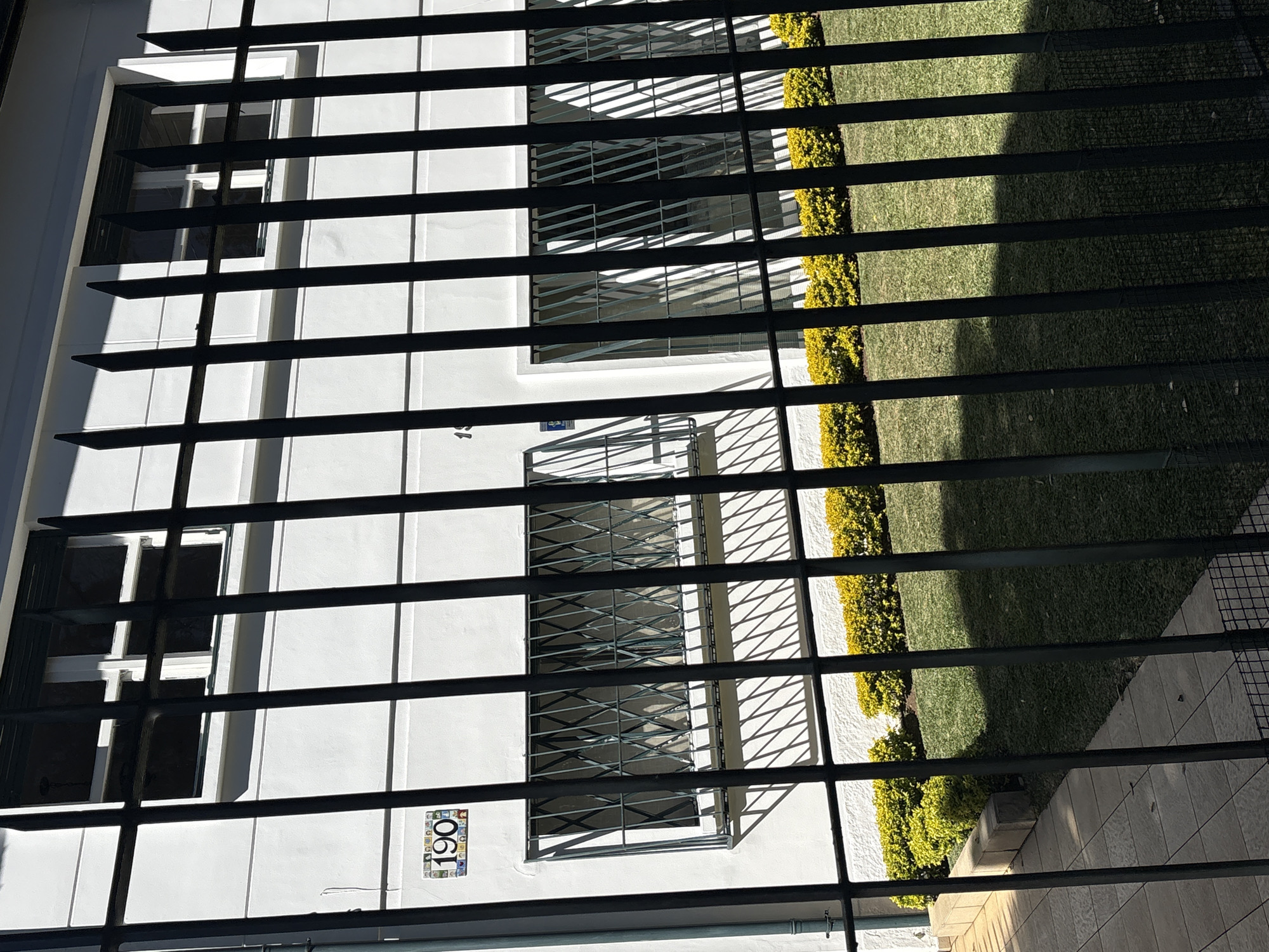The image shows a white building with windows and a decorative tile mosaic above one of the windows that reads "190". The building is behind a black metal fence. A well-maintained lawn with a yellow bush border is in front of the building. The scene appears to be a residential or commercial property in a city. The building facade is clean and white, with modern window frames and bars. The ground is paved with light-colored tiles on the left and covered with green grass on the right. The lighting suggests it is a sunny day with shadows cast by the fence and building. The overall impression is one of order and neatness.