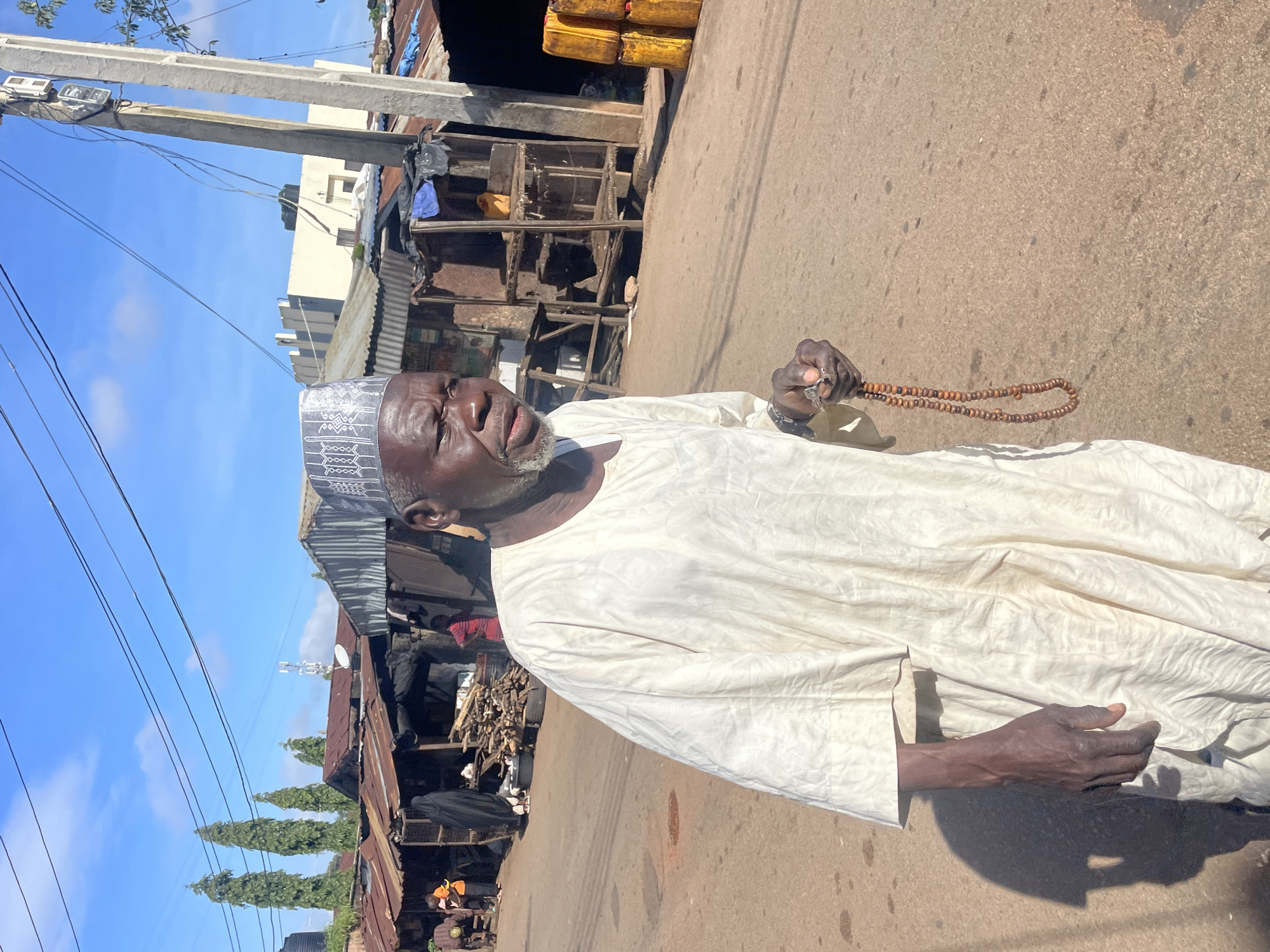 An elderly Black man, wearing a white traditional garment and a patterned cap, stands on a dirt road in what appears to be a market area in Jos, Nigeria. He is holding a string of brown prayer beads in his right hand. The background features several small, makeshift stalls with corrugated metal roofs, and some stacked yellow containers. A utility pole with electrical equipment stands to the right of the man. The sky is clear and blue with some clouds.