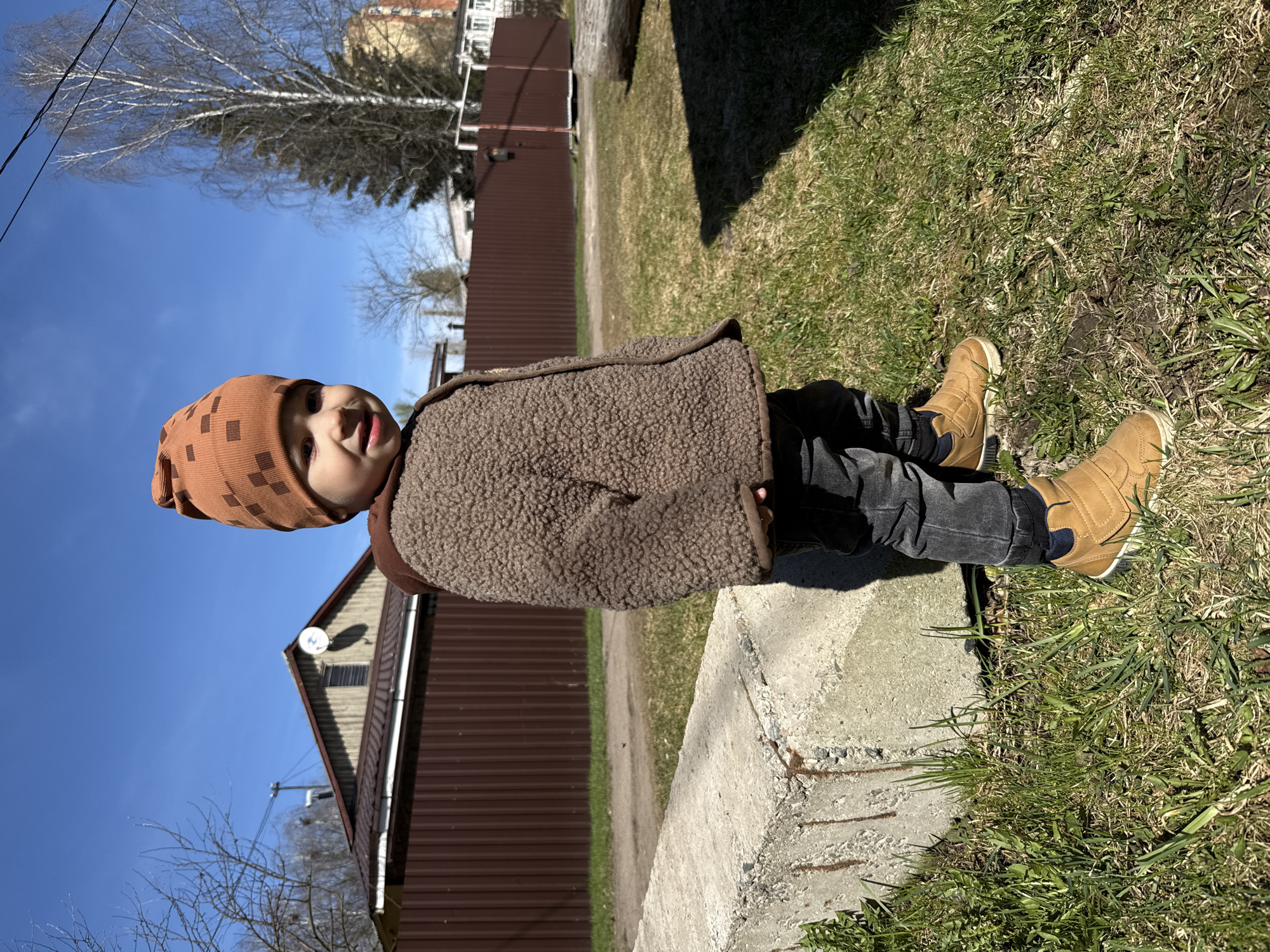 A young child, likely a toddler, is depicted sitting upright on a concrete block outdoors. The child wears a patterned brown beanie, a textured brown fleece jacket, dark trousers, and tan high-top boots. They are looking towards the camera with a slightly open mouth and a faint smile, conveying a sense of curiosity or happiness.
The immediate surroundings feature green and dry grass beside the concrete block. In the mid-ground, a long, brown corrugated metal fence stretches horizontally. Beyond the fence, a house with a matching brown roof and a white satellite dish on its side is visible. Bare trees, suggesting early spring or late autumn, stand against a clear, vibrant blue sky. Overhead, power lines traverse the upper left portion of the frame. The scene is brightly illuminated by natural sunlight, indicating it is daytime. There is no discernible text within the image. The setting appears to be a residential or semi-rural area in Bor, Russia.
