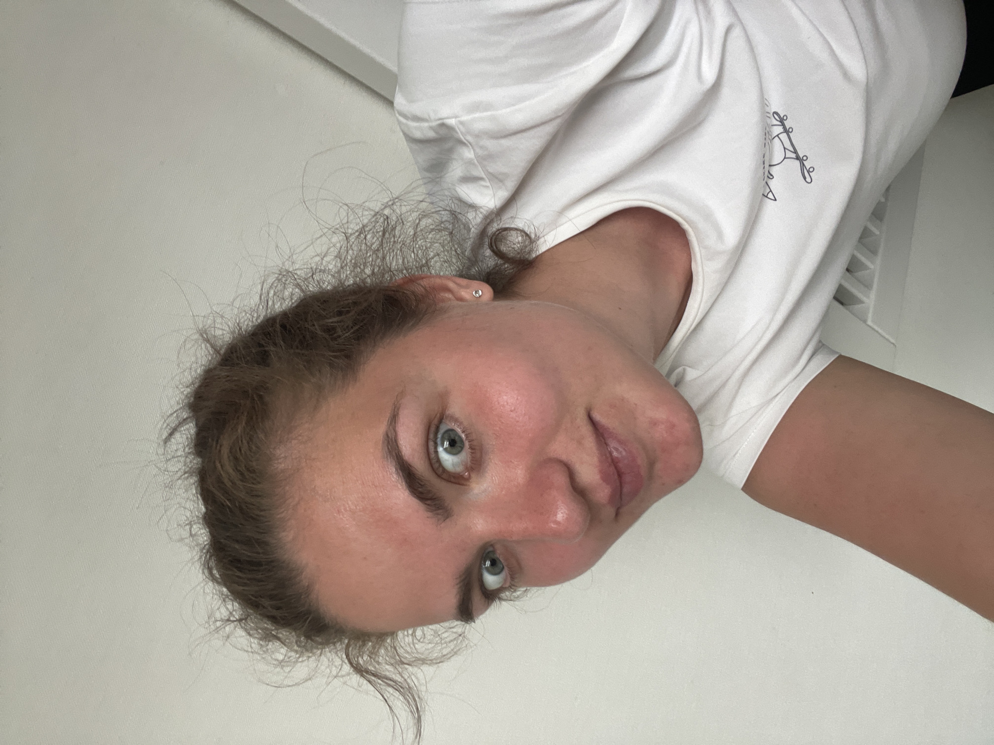 A young woman with light skin, light brown curly hair, and blue eyes is visible from a low angle. She's wearing a white t-shirt with a small graphic on the chest. The background appears to be a plain, light-colored wall and a vent. No specific location in Moscow is discernible.