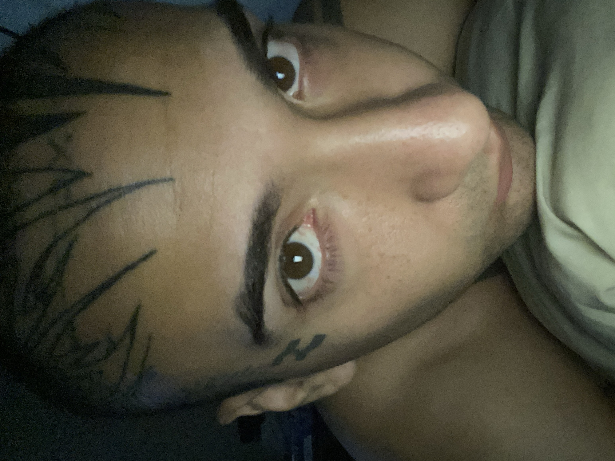 A close-up, portrait-oriented image depicts a person's face. The individual has brown eyes and dark hair, with what appear to be tattoos on the face and head. One visible tattoo is located near the temple, and another smaller one is under the eye. The skin tone is medium. The person is positioned at an angle, looking upwards and slightly to the side. Part of the torso and what appears to be a pillow or bedding are visible. The environment is dimly lit. There is no indication of location of Buenos Aires, Argentina.