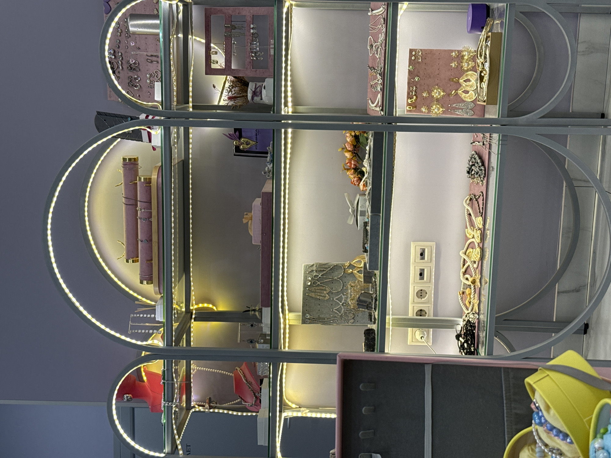 The image shows a well-lit display case filled with jewelry. The display case is made of glass shelves and metal frames, with LED strip lights illuminating each shelf. On the top shelf, there are pink jewelry holders with necklaces. The second shelf has more jewelry, including necklaces and bracelets, displayed on a pink felt mat. The third shelf features a variety of earrings displayed on a grey board, along with other jewelry items. The bottom shelf has several necklaces laid out on a pink mat. The overall impression is that of a jewelry store or a display within a retail environment. The image appears to be taken indoors in a retail setting. The location is specified as Lyubertsy, Russia, suggesting this is a commercial establishment in that city. There are no people visible in the image.