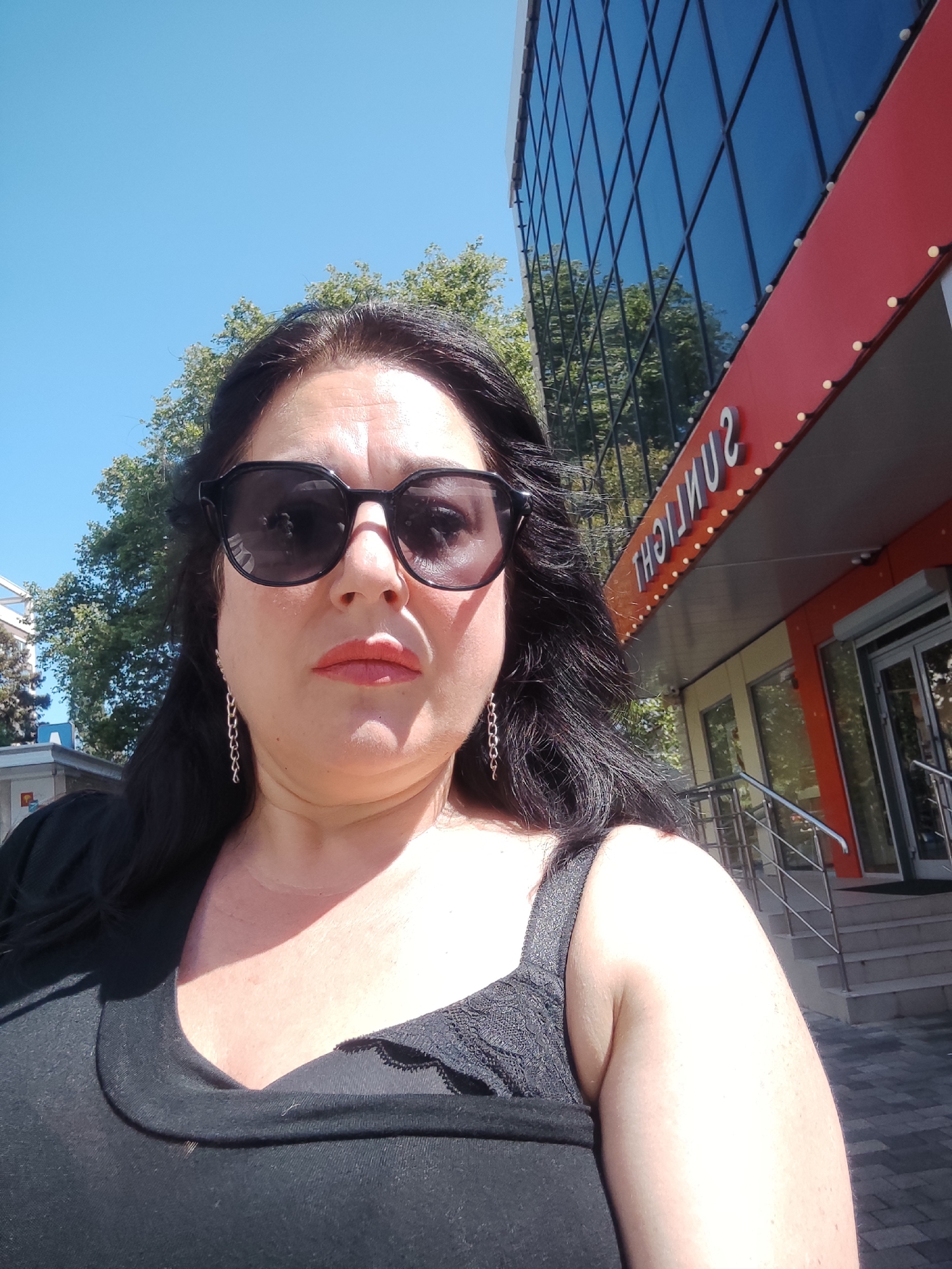 A woman is taking a selfie in front of a building in Sochi, Russia. She is wearing sunglasses and a black top, and has long, dark hair. The building is modern with glass windows and a red facade. The word "SUNLIGHT" is visible on the building in illuminated letters. The sky is blue and there are trees in the background. The woman is close to the camera and appears to be outdoors.