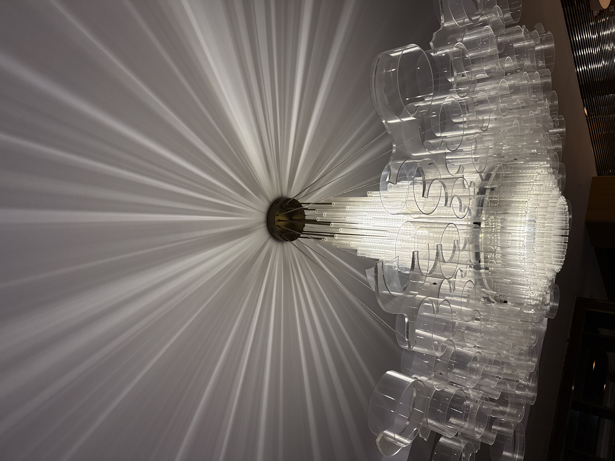 The image shows a modern chandelier hanging from a white ceiling, casting dramatic, radial shadows. The chandelier is made of clear, cylindrical glass or acrylic tubes of varying lengths, arranged in layers. The central part of the chandelier is illuminated, and the light diffuses through the tubes, creating a sparkling effect. The shadows projected onto the ceiling are sharp and distinct, resembling sunbeams or the spokes of a wheel. The image is taken from a low angle, looking upwards. The lower portion of the image shows a dark wooden frame, possibly part of a doorway or window, and a slatted surface that could be a ventilation grate or part of a decorative element. This chandelier and its lighting effect are likely located in an interior space in Bodrum, Turkey, possibly a hotel lobby, restaurant, or a high-end residence. There are no people visible in the image.