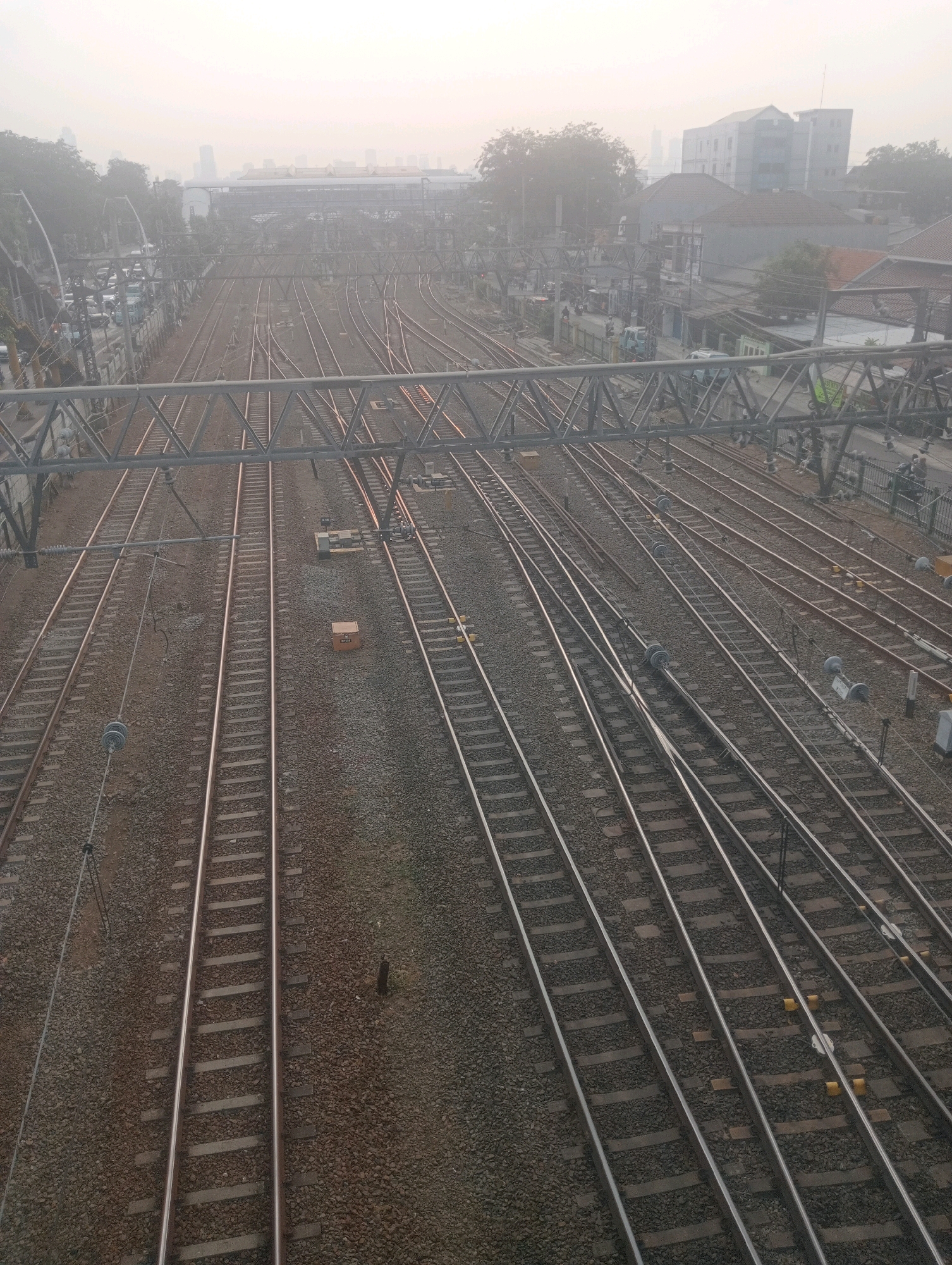 The image captures an aerial view of a dense network of train tracks in East Jakarta, Indonesia. The scene is enveloped in a hazy atmosphere, suggesting smog or fog, which partially obscures the urban landscape in the background. Numerous parallel and intersecting train tracks dominate the foreground, laid on a ballast of small stones. Overhead electrical lines and their support structures are visible, spanning across the tracks. A pedestrian overpass with a metal truss structure is present in the upper portion of the image. In the mid-ground, buildings of varying heights and architectural styles can be seen, some appearing to be residential, while others seem to be commercial or industrial. Further in the distance, taller buildings suggest the presence of a more developed urban center. The overall impression is one of a busy transportation hub within a sprawling metropolitan area, characterized by a high density of infrastructure and a subdued, muted color palette due to the atmospheric conditions.