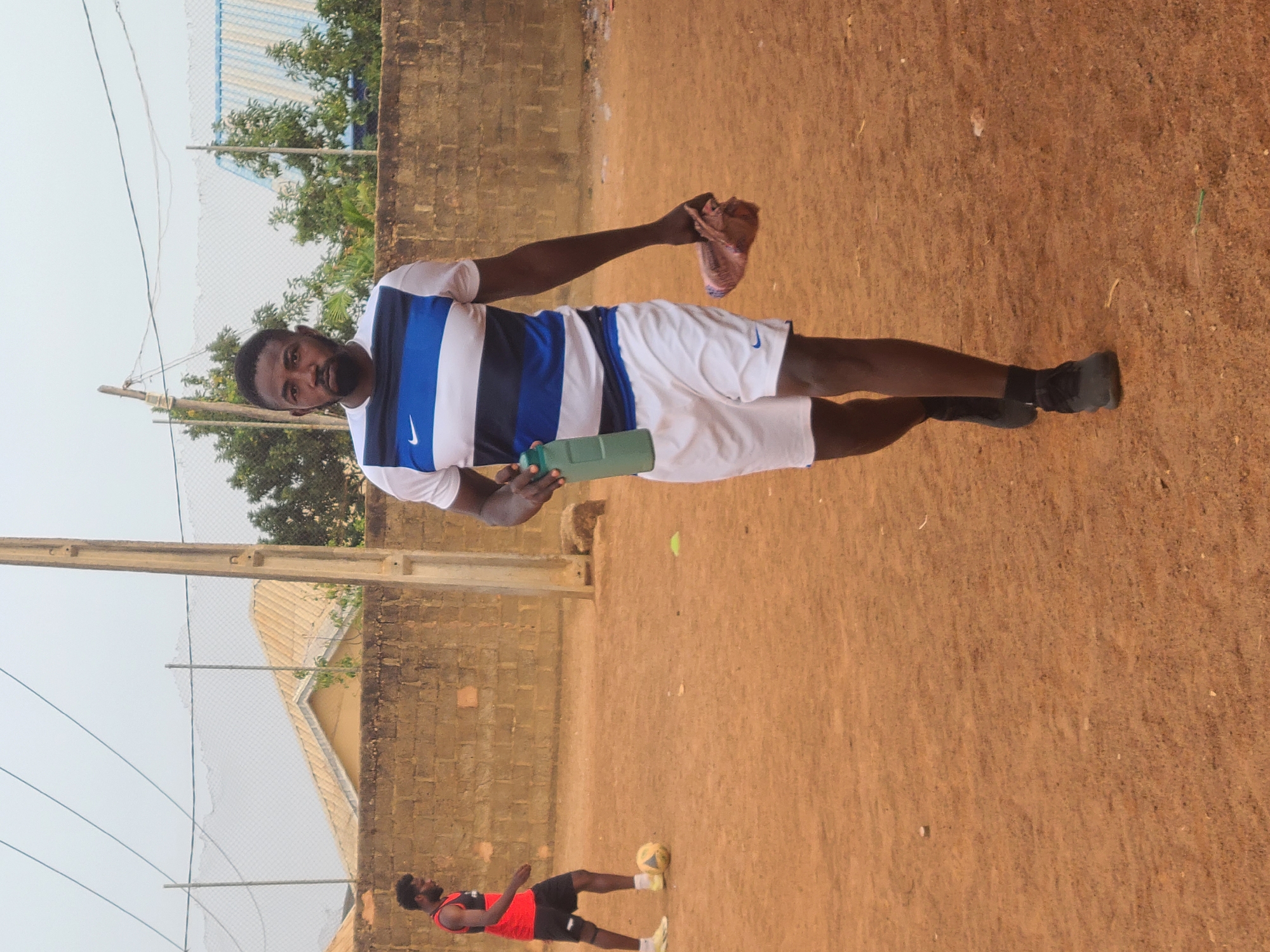A medium shot depicts an adult male standing on an ocher-colored, earthen surface in Barakin Lerawi, Nigeria. He wears a white and blue striped t-shirt, white shorts, and dark-colored shoes. He holds a green container in one hand and a brown object, possibly a sock, in the other. In the lower left corner, another male figure, smaller in scale due to distance, is seen kicking a soccer ball on the same surface. A low brick wall runs along the background, with trees and a metal structure visible above it. Electrical wires and poles are present in the left background.