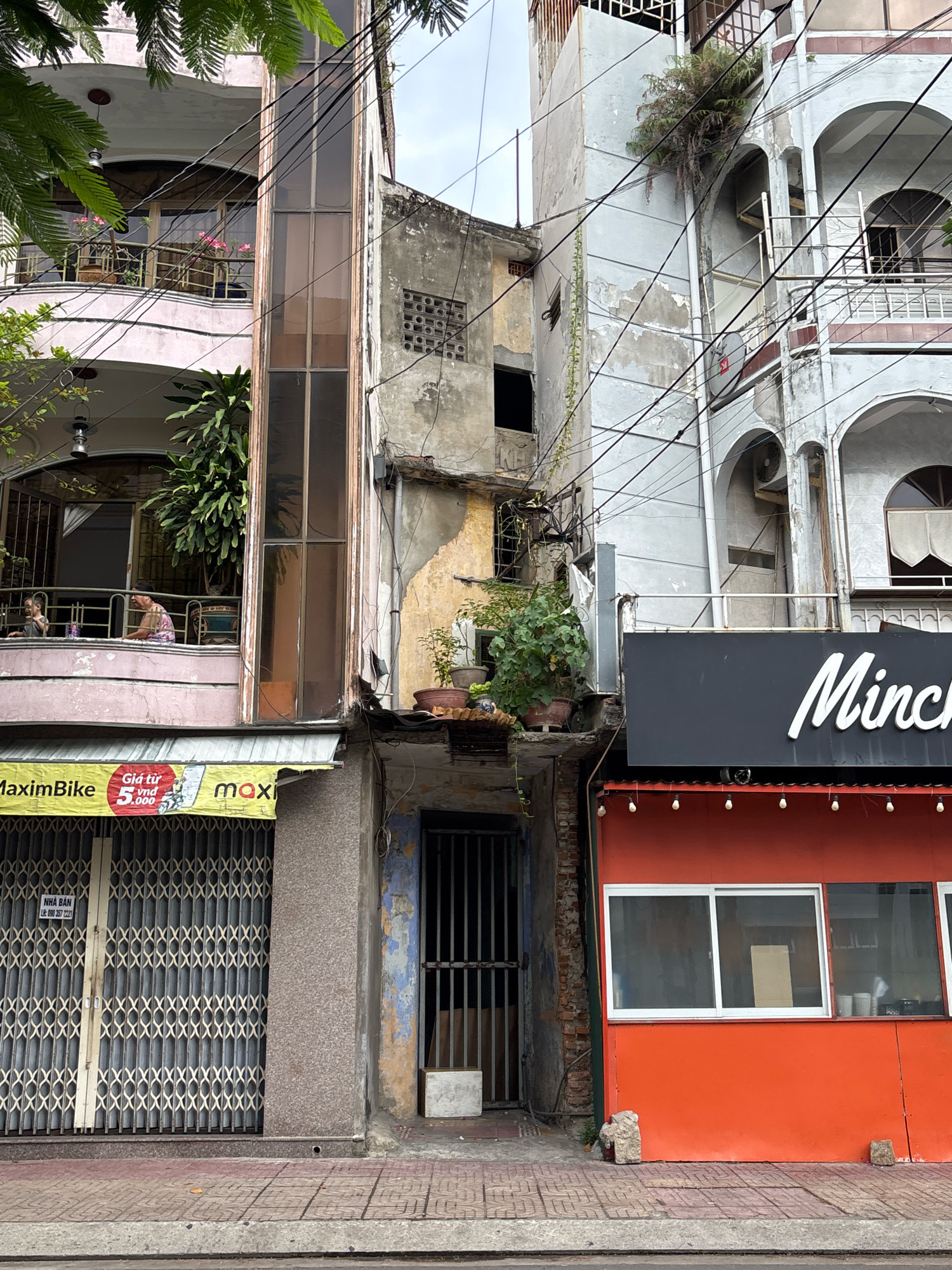 This is a street-level view of an urban scene in Nha Trang, Vietnam, likely on a cloudy day. The image captures a section of buildings with varying architectural styles and states of repair.
On the left, a pink multi-story building features balconies with railings and potted plants, including a large leafy plant on an upper balcony. A person is visible on one of the balconies. Adjacent to this building is a glass-enclosed structure with vertical frames. Below these structures, a yellow awning with text and graphics, and a closed metal gate with a patterned design are visible. Another sign with text is also present above the gate.
To the right of the pink building is a much older, weathered building with exposed brickwork and peeling paint. This building has a narrow, enclosed walkway or stairwell with a metal grate door. Potted plants are also present on ledges of this structure.
Further to the right, a more modern, light-colored building with arched openings and balconies is visible, partially obscured by electrical wires. A dark gray sign with white script reading "Minch" is mounted above an orange-fronted structure with a white-framed window. The orange structure has a row of small lights along its upper edge.
The ground in front of the buildings is paved with patterned tiles. The scene suggests a densely populated urban environment with a mix of old and new construction, and evidence of overhead electrical wiring crisscrossing the scene.