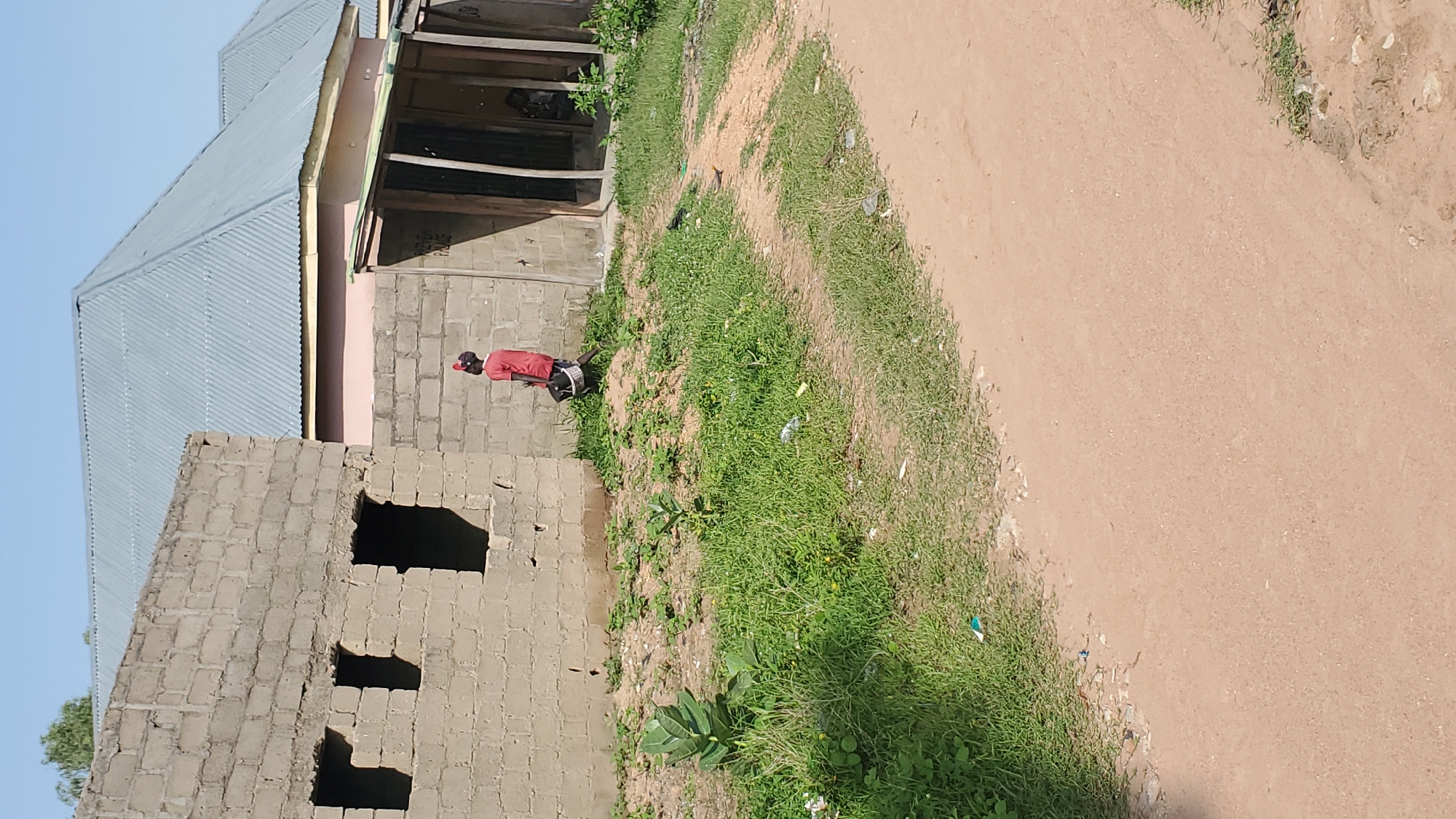 A person, possibly female, wearing a red shirt, dark shorts, and a red headscarf, walks on a dirt path next to a partially constructed building. The person is carrying a white bucket in their left hand. The building is made of grey concrete blocks with unfinished openings for windows. Behind this building, a completed structure with a grey corrugated metal roof is visible. Green grass and some scattered debris are present on the ground. The scene appears to be in an outdoor, semi-urban environment, likely in Jalingo, Nigeria, given the architectural style and vegetation. The sky is clear and blue.