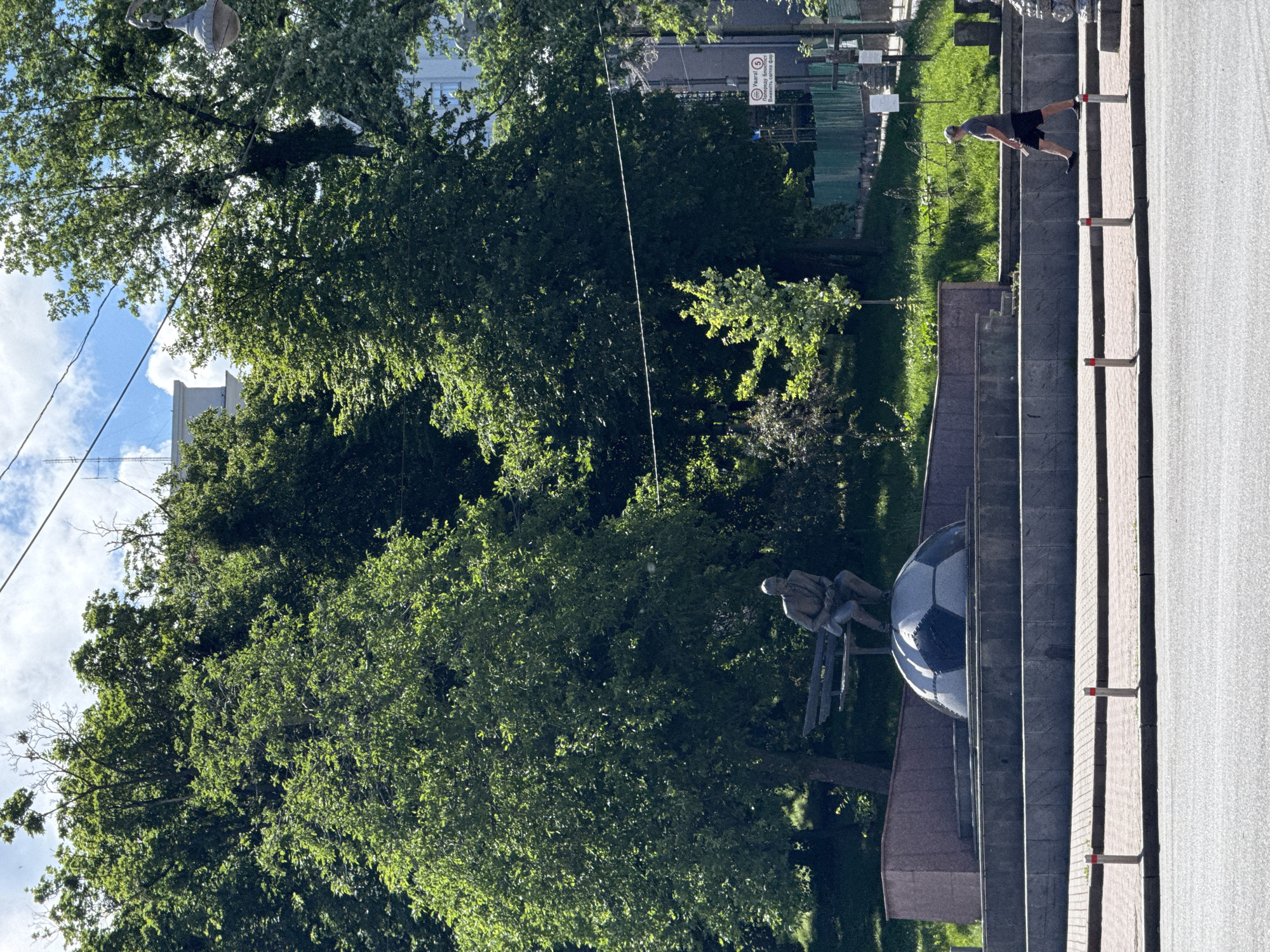 In Kyiv, Ukraine, a man walks past a monument depicting a statue of a seated man on a large soccer ball, surrounded by lush green trees and a cloudy sky. The monument is situated on a tiered stone base. In the background, a building and street signs are visible. A street light hangs from a pole to the right of the frame. The man is wearing a grey t-shirt, black shorts, and a baseball cap. He is holding a white object in his right hand. The man is walking from right to left, away from the monument. The scene is captured on a sunny day, with dappled light illuminating the monument and the surrounding area.