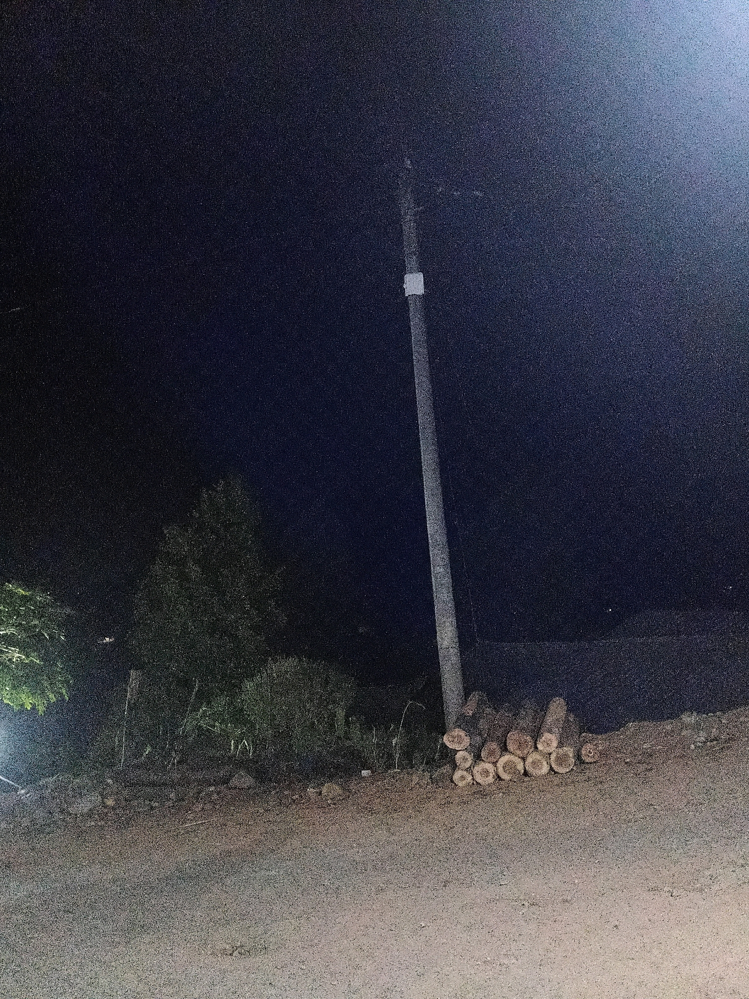 The image depicts a nighttime scene in Sinduaji, Indonesia. A single, tall, slender utility pole stands vertically near the center of the frame, with a small white rectangular marking about a third of the way up its height. To the right of the pole, a pile of chopped logs rests on the ground. The logs are cut into varying lengths, and their circular ends are visible, revealing their grain. The ground in the foreground is unpaved and appears to be a dirt or gravel path. In the background, to the left of the pole, are several trees and dense vegetation, some of which are illuminated by a light source, casting faint shadows. The sky is dark, indicating nighttime, and the overall lighting suggests the presence of artificial illumination, possibly from the utility pole itself or another nearby light source. There are no people or animals visible in the image. The scene appears to be an outdoor, rural or semi-rural area.
