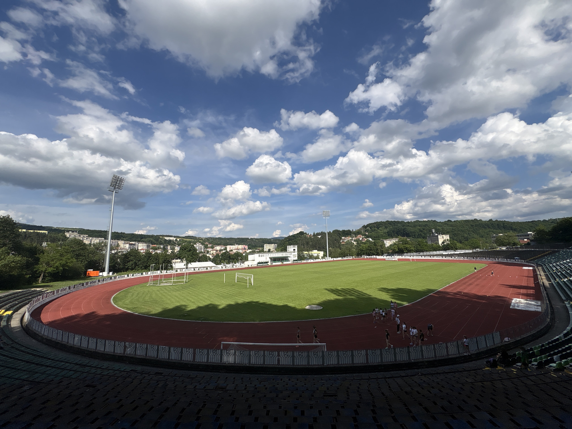 An outdoor athletic stadium is shown under a bright blue sky with scattered white clouds. The stadium features a red running track surrounding a green grass field, which is marked with white lines and includes two soccer goals. Stadium seating, partially in shadow, is visible in the foreground and along the right side. Several people, appearing to be athletes, are scattered on the track and field. In the background, a city with buildings and trees is nestled on rolling hills. The stadium lights are tall and prominent. The stadium is located in Gabrovo, Bulgaria.