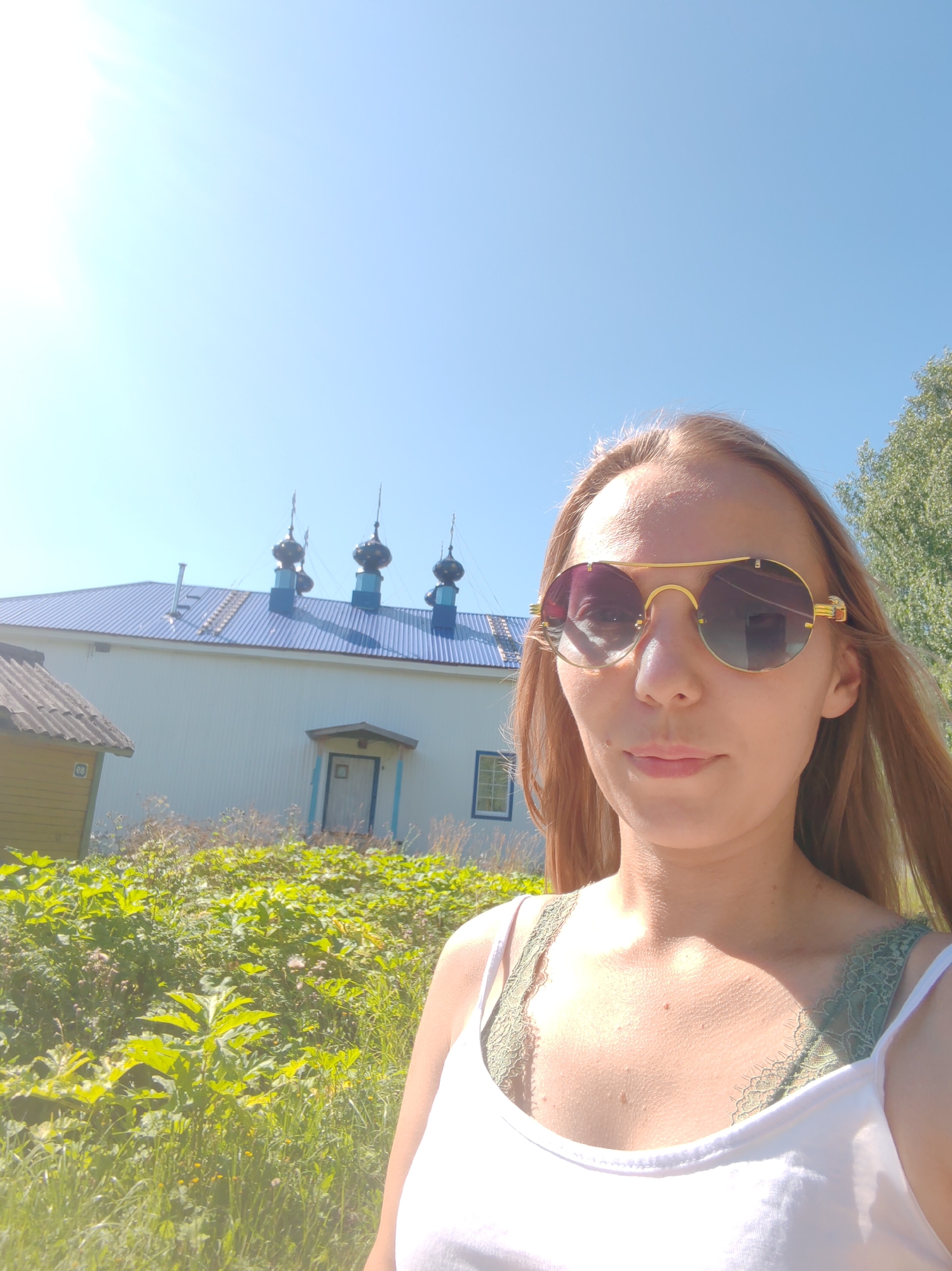 A medium shot shows a young woman with blonde hair wearing sunglasses and a white tank top with a green lace bra underneath. She is outdoors, with a bright blue sky above and a building with a blue roof and cupolas in the background. To the left of the building, a small yellow structure is visible. Lush green foliage and grass are in the foreground. The image is taken outdoors on a sunny day.