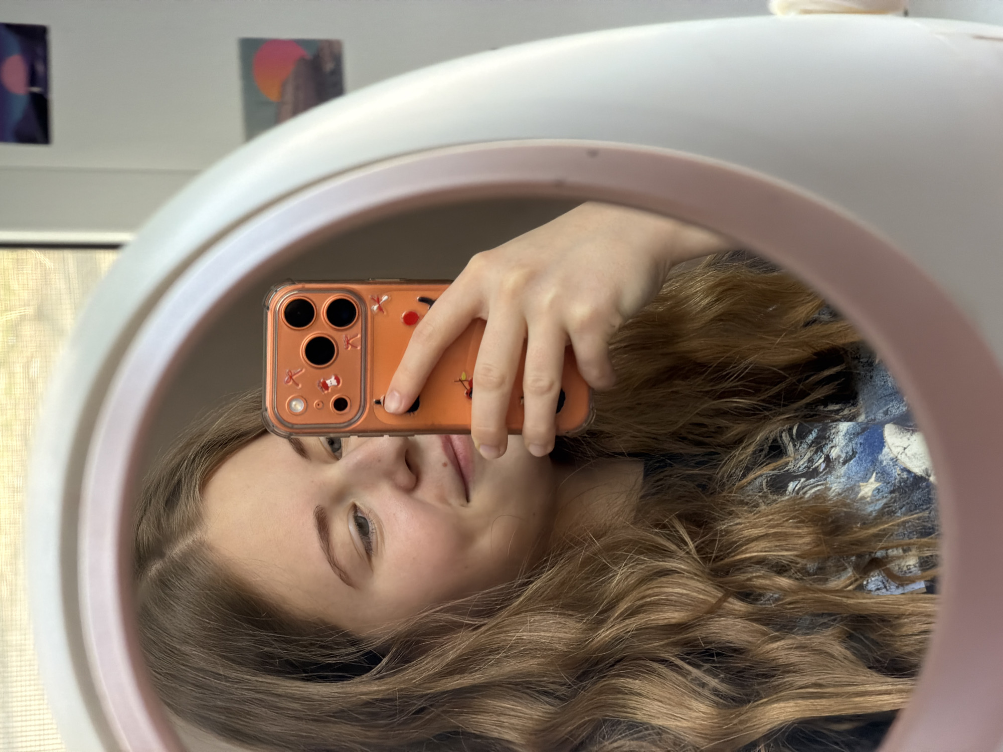 A selfie in a circular mirror with a light ring. A person is holding a phone, with an orange case, up to the mirror. The person has light skin, long brown hair, and is smiling softly. The mirror is positioned in front of a wall with two posters hanging on it. The location of Perm, Russia is not discernible in the image.