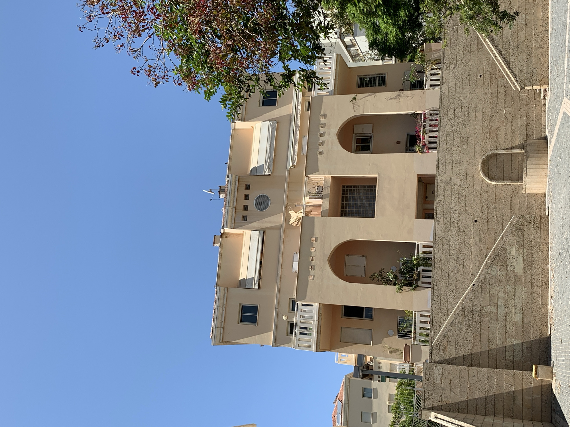 A vertical shot of an apartment building in Tel Aviv, Israel. The building is cream-colored and has multiple floors. There are several windows and balconies visible, some of which are decorated with plants. The building is surrounded by trees and other vegetation. The sky is blue and clear. The foreground of the image shows a sidewalk and part of a stone wall.