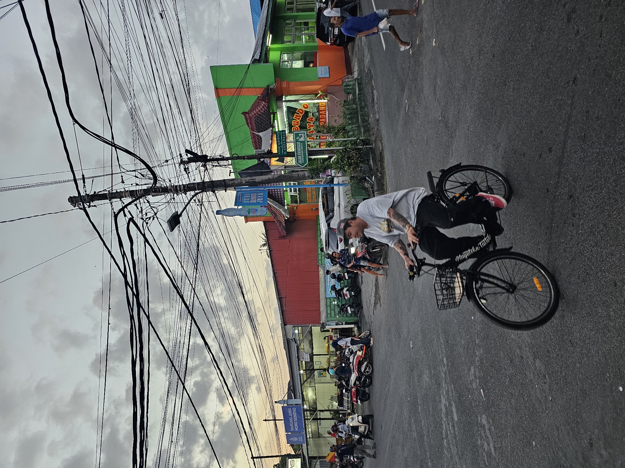 The image captures a street scene in Denpasar, Indonesia, likely during the late afternoon or early evening, given the overcast sky with hints of sunset colors. The foreground features a person riding a bicycle, wearing a grey t-shirt and black pants, with a basket attached to the handlebars. The bicycle's front wheel is slightly blurred, indicating motion.
In the mid-ground, the street is busy with several motorcycles parked and people standing around. A building with a red facade is visible, and further to the right, a building with a bright green and orange exterior is prominent. This green and orange building has signage indicating it is "Bundo Jaya" and mentions "Jl. Sudamala" and "Jalur Evakuasi" (Evacuation Route), suggesting it might be a restaurant or establishment. Another establishment to the left displays a sign for "WARUNG JAWA MORO SENENG."
The background is dominated by a dense network of overhead electrical wires and poles, a common sight in urban Indonesian settings. A few vehicles, including cars and more motorcycles, are present on the street. A person in a blue t-shirt and shorts is walking on the right side of the road, carrying a white object. The overall impression is a bustling urban environment with a mix of pedestrian and vehicular activity, overlaid with extensive utility infrastructure.