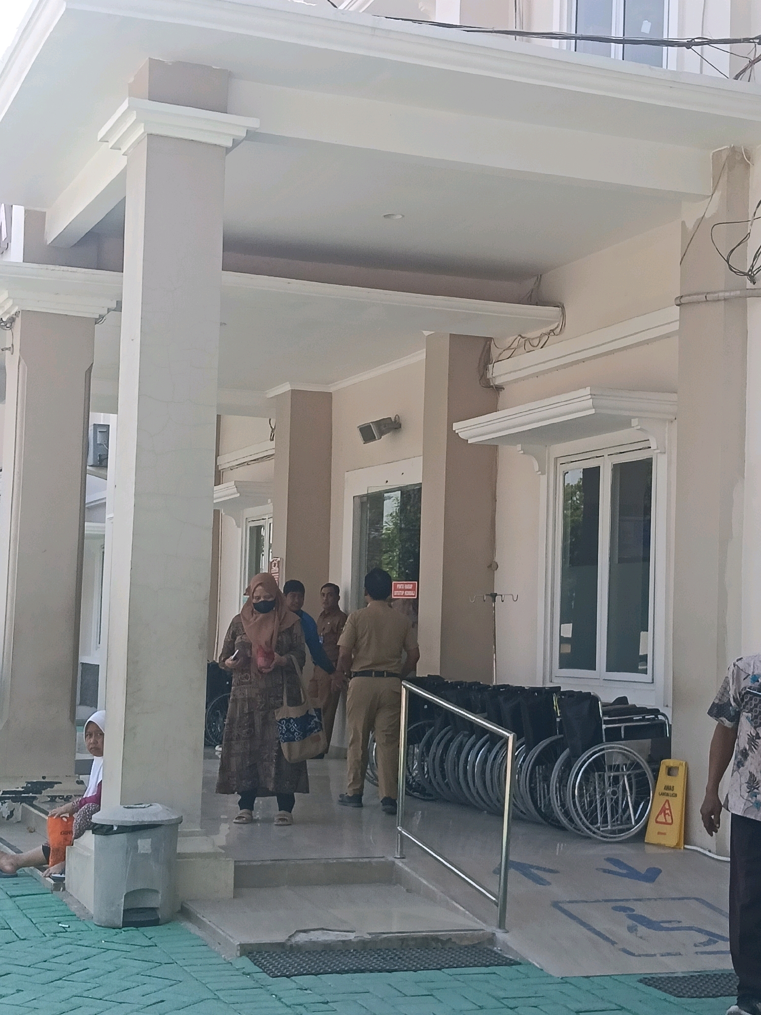 This image depicts the exterior of a building in Balun, Indonesia, with a covered walkway and an entrance. In the foreground, several wheelchairs are neatly lined up, some folded, near a ramp with a blue wheelchair accessibility symbol painted on the ground. A yellow sign with a warning symbol is also visible next to the wheelchairs. Several people are present in the walkway. A woman wearing a hijab and a patterned dress is walking towards the left, carrying a bag. Two men in uniform, possibly staff members, are standing near her, and another man in uniform is seen in the background. To the left, a young girl wearing a white headscarf and a colored outfit is sitting on a low platform next to a grey trash can. The building has large windows and beige-colored walls, with prominent white pillars supporting the roof of the walkway. The ground outside the walkway is paved with green tiles arranged in a pattern.
