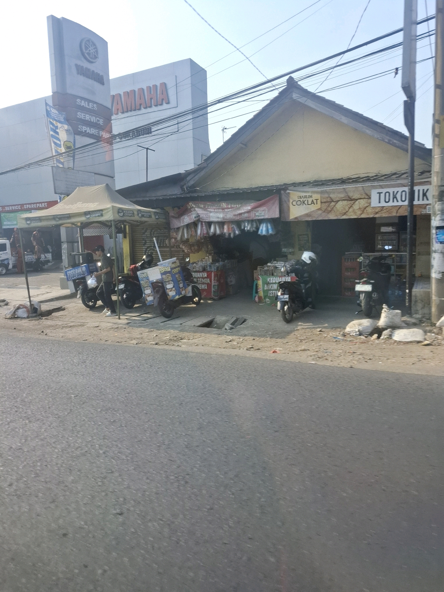 The image depicts a street scene in Karangtengah, Indonesia, featuring a Yamaha dealership and a small shop. In the foreground, a paved road is visible. To the left, a large Yamaha building with prominent signage stands. A tall signpost with "SALES", "SERVICE", and "SPARE PART" is present. Adjacent to the building, a white van is parked. In front of the shop, there is a tented awning under which several food vendors operate. Two motorcycles, one with a blue and white advertising box on the back, are parked near the shop entrance. A person in a dark shirt and black pants is standing next to one of the motorcycles. The shop itself has a light-colored facade and appears to sell various goods, with items displayed both inside and outside. Signs in Indonesian, such as "DIARJUM COKLAT" (Chocolate Advertisement) and "TOKO IK" (likely part of "TOKO IKAN" or fish shop, or a general store name), are visible on the shop. The environment is outdoors, with a clear sky and overhead electrical wires present. Piles of white sacks or debris are located on the ground near the shop.