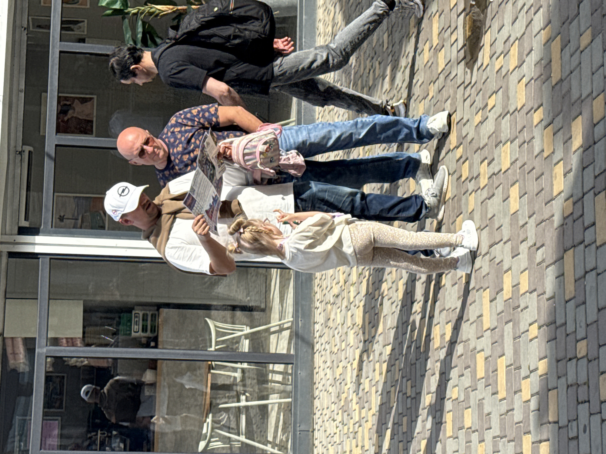 The image shows a group of people standing on a brick paved street in front of a building with large glass windows. The first man is wearing a white t-shirt, beige sweater, and white baseball cap, holding a newspaper. A young girl wearing a white shirt and patterned leggings stands to his right. Behind them, a bald man in a blue patterned shirt is reading the newspaper with the first man. A pink children's backpack rests on the shoulders of the bald man. Behind the bald man, a man wearing a black t-shirt, gray jeans, and a black backpack is walking. The building behind them is reflected in the glass, showing a person sitting at a table. The location is in a city of Unknown, Unknown.