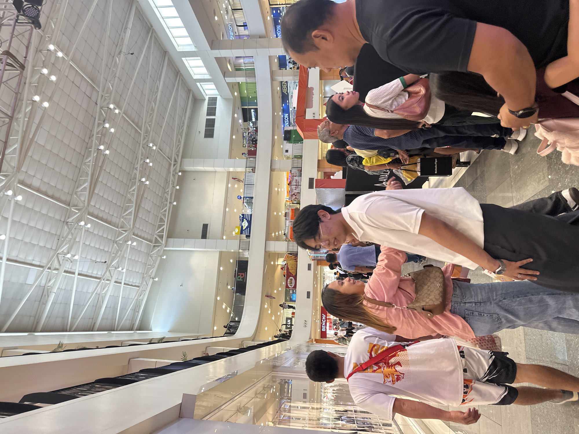 The image depicts an indoor scene within a multi-level shopping mall. Several individuals are visible, appearing to be in a queue or moving through a common area. The architecture suggests a modern commercial space with a high, exposed ceiling structure featuring numerous lights and ventilation systems. Balconies or upper levels with railings overlook the main floor, where shops are visible in the background, some with illuminated signs. The floor is tiled, and the overall lighting is bright, characteristic of a well-lit mall environment. The presence of various shops and the general atmosphere indicate a public shopping or commercial district. This scene is consistent with locations such as SM Lanang Premier, located in Davao City, Philippines, a prominent shopping mall in the region.