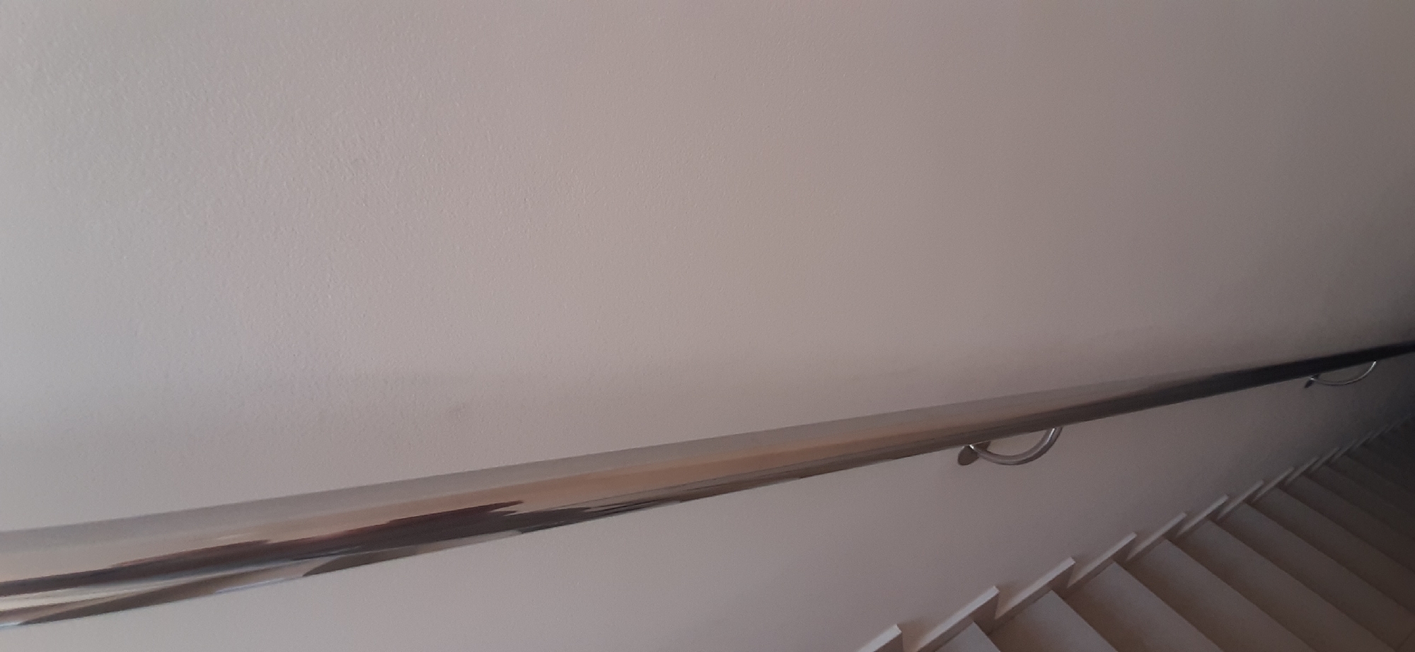This image shows a close-up view of a modern staircase with a sleek metal handrail. The handrail is polished chrome and extends diagonally across the frame, supported by discreet brackets. Below the handrail, the white steps of the staircase are visible, receding into shadow. The wall adjacent to the handrail is a neutral, pale color, showing a slight texture. There are no people or animals present in the image. The lighting is soft and even, suggesting an indoor environment, likely a home or public building. The overall impression is one of clean lines and contemporary design. The image does not provide any specific details about its location in the city of Yessentuki, Russia.