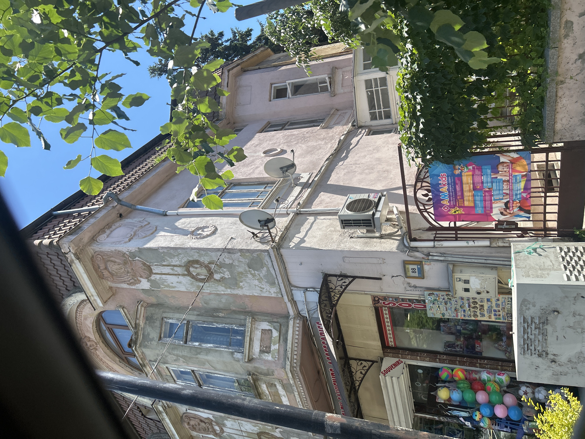 The image shows a building on a city street, identified as being in Varna, Bulgaria. The building appears to be old, with peeling paint and ornate architectural details. The lower part of the building houses shops, one of which has a sign that reads "SOUVENIRS". In front of the shops, there are displays of colorful balls. A metal gate with a colorful advertisement for "AMAKids" is visible to the right. Lush green foliage and trees are present in the foreground and against the bright blue sky. The scene suggests a sunny day in an urban environment.