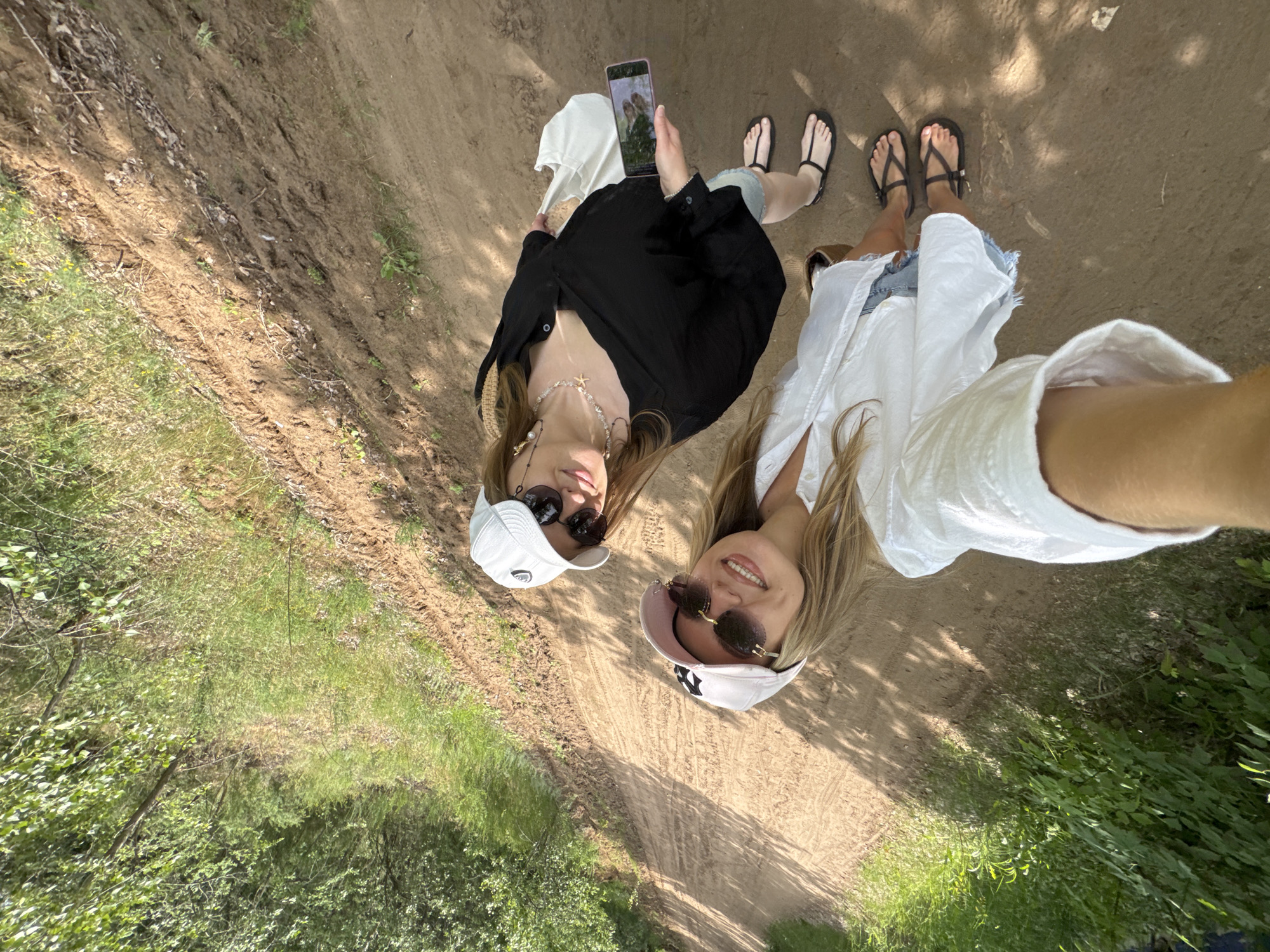 Two women are posing for a selfie on a dirt path in a wooded area. The woman in the foreground is wearing a white baseball cap, sunglasses, and a white open-collar shirt. The woman behind her is wearing a white baseball cap, sunglasses, a black collared shirt, and denim shorts. She is holding a smartphone in her right hand. The path is surrounded by trees and vegetation. The lighting suggests it is daytime.