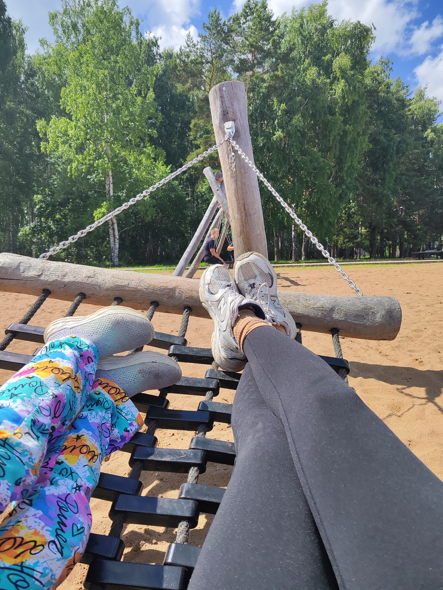 In an outdoor playground, two individuals are seated on a black rope and wooden beam swing, their legs crossed. One person wears colorful, patterned leggings and light-colored sneakers, while the other wears black leggings and grey sneakers. In the background, a person sits on a swing further back. The playground is situated in a sandy area with a backdrop of lush green trees under a partly cloudy blue sky. The scene is located in Chaikovsky, Russia.