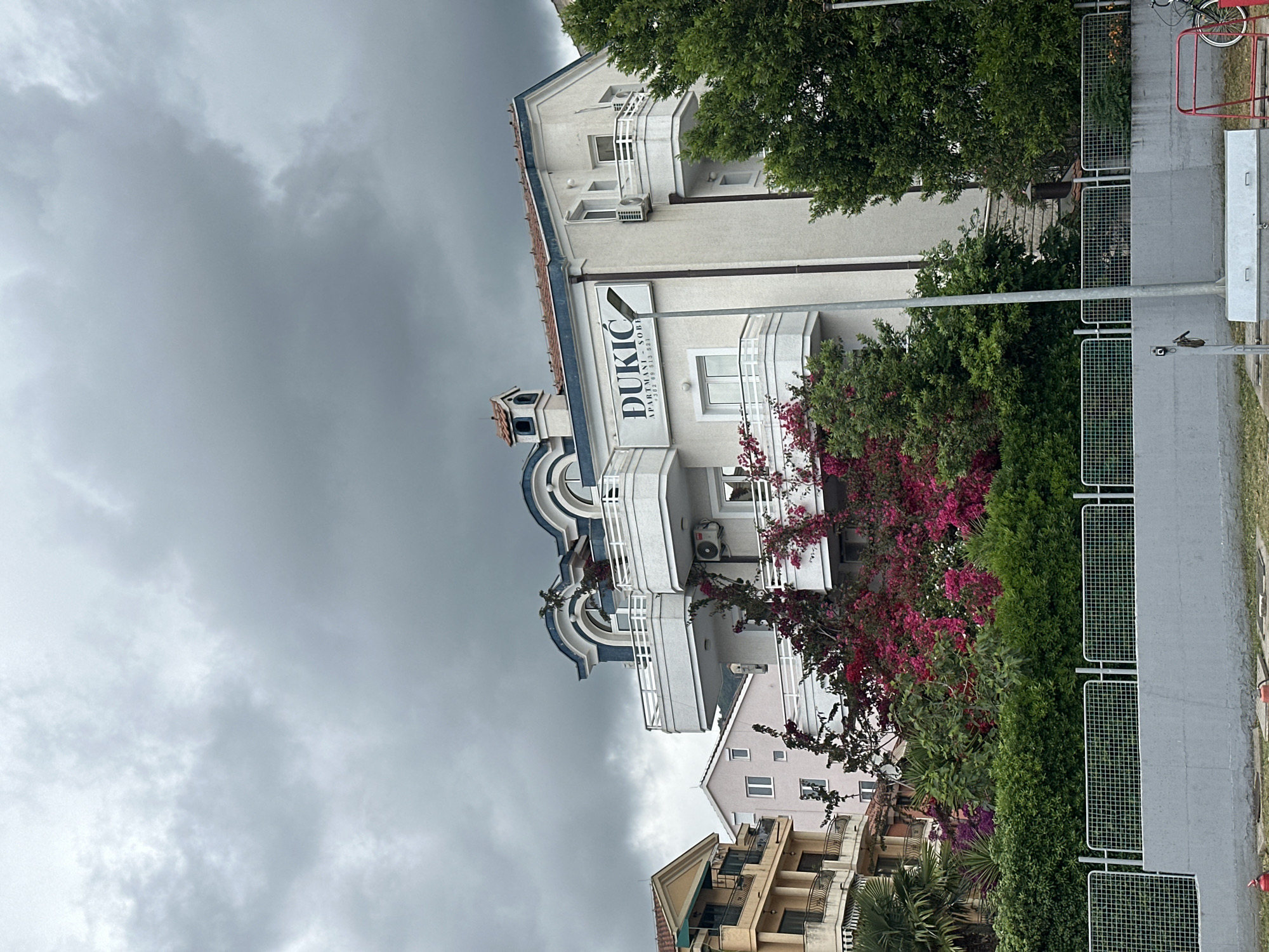 The image shows a building in Budva, Montenegro, under a cloudy sky. The building, named "Dukic," has a white facade with multiple balconies and a sign with its name. The building is surrounded by lush greenery, including flowering plants with vibrant pink blossoms. The image is taken from a slightly low angle, emphasizing the building's height and presence. A sidewalk with a metal fence runs along the right side of the image. Other buildings are visible in the background, suggesting a densely populated urban environment. A street lamp can be seen on the right. A bicycle is parked beyond the metal fence. The atmosphere appears overcast and potentially damp.