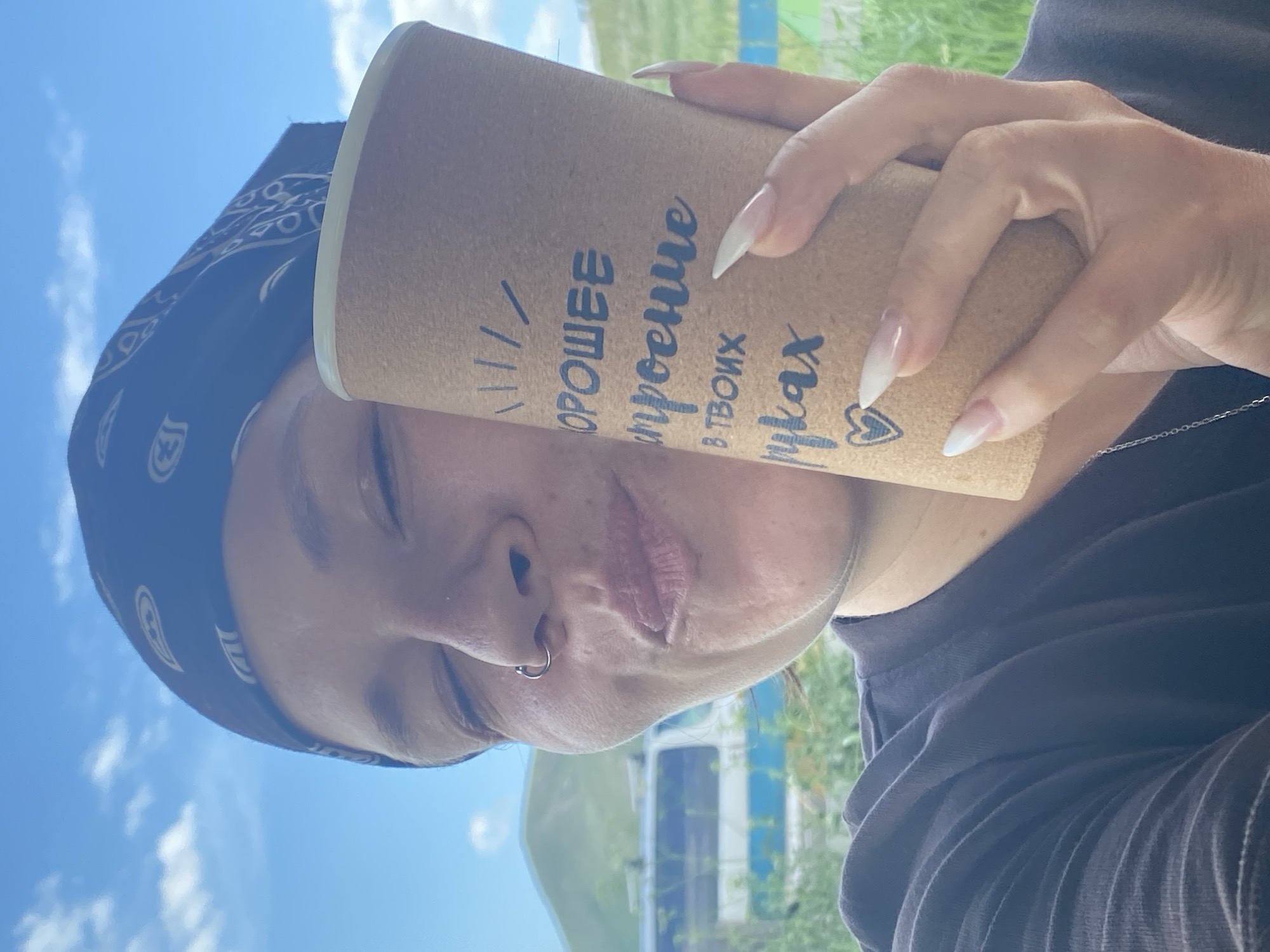 In Абзаново, Russia, a close-up, selfie-style image captures a person holding a reusable cup. The person, identified as female by her appearance and long, manicured nails, wears a dark t-shirt and a black bandana with a white paisley pattern. She is looking towards the camera with her eyes closed and her lips puckered, suggesting a playful or affectionate gesture. A silver nose ring is visible on her left nostril. The cup, made of a light brown, textured material, displays text in a dark font, partially obscured by the person's hand. The background features a bright blue sky with scattered clouds, a green, rolling hill, and a blurred vehicle. The overall lighting suggests a sunny day.