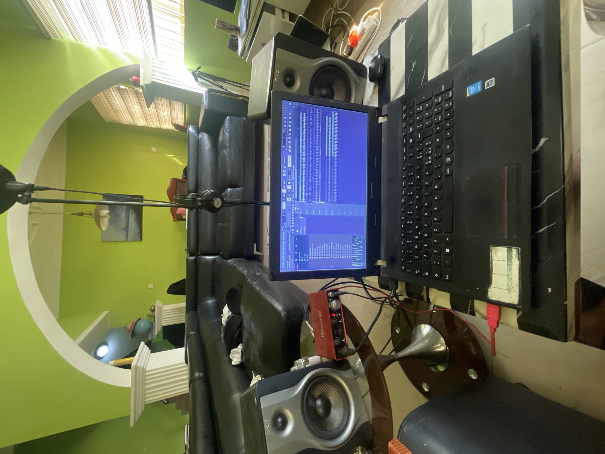 The image displays an indoor home studio or music production setup, with the original photo appearing to be rotated 90 degrees clockwise. The setting is a room with vibrant lime green walls and a distinctive white architectural archway. Bright natural light streams in through venetian blinds, indicating daytime.
At the forefront, a black Lenovo laptop is open, its screen displaying a Digital Audio Workstation (DAW) interface with multiple audio tracks and waveforms, suggesting active music production or editing. A red USB drive is plugged into the laptop, and a black computer mouse rests nearby on a black and white striped tabletop.
A red Focusrite Scarlett audio interface is prominently placed next to the laptop, connected by various cables. Below it, a grey studio monitor speaker, bearing the text "RXE" or "RVE," sits on a table with a round wooden base and a metallic stand. Another matching studio monitor is partially visible behind the laptop.
To the left, a microphone stand with a shock mount is positioned, ready for vocal or instrument recording. A black leather-like sofa or couch fills the mid-ground, providing seating. In the background, a small globe rests on a white column-like pedestal, and a framed picture adorns the wall. The visible text includes "Lenovo," "Focusrite," "RXE" (or "RVE"), and a Windows logo sticker on the laptop. The overall scene depicts a dedicated and equipped space for creative audio work, situated in Abuja, Nigeria.