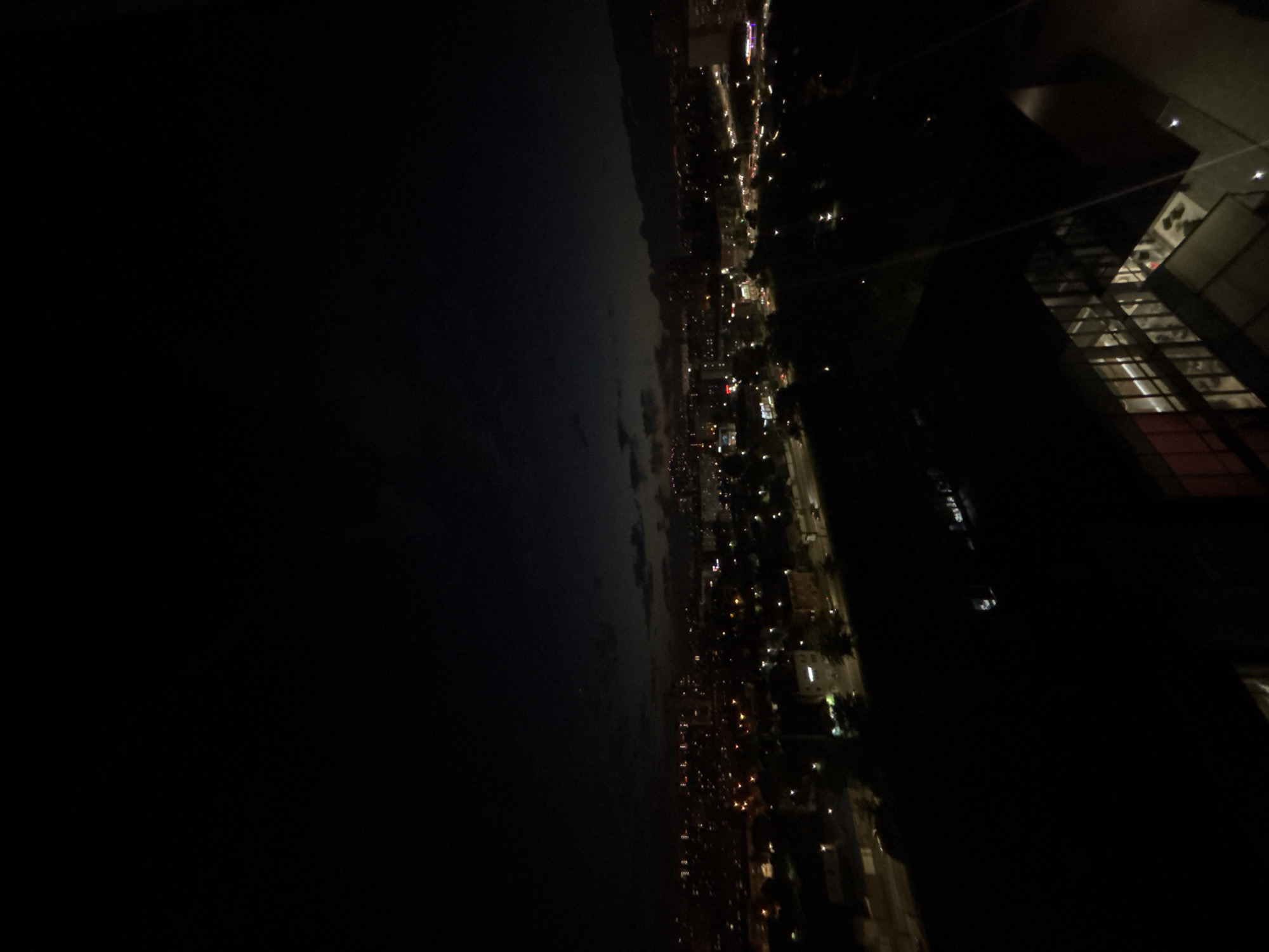 The image is a nighttime aerial view of a city, possibly Krasnodar, Russia. The scene is dominated by darkness, with the city lights providing the only illumination. Buildings and streetlights are visible, forming a pattern of illuminated dots against the blackness. The lights suggest a populated urban area with ongoing activity. The perspective is from a high vantage point, looking down and across the cityscape. To the right, a modern building with illuminated interior spaces is partially visible, indicating the presence of architecture that is lit from within. The overall impression is one of urban sprawl at night, with the vastness of the dark sky and land emphasizing the scattered nature of the city lights. The weather appears clear, as there are no visible atmospheric obstructions like fog or heavy clouds obscuring the view. The time of day is clearly night, suggested by the darkness and the artificial lighting. No people or specific objects are clearly discernible, beyond the general elements of a city at night. There is no visible text in the image.