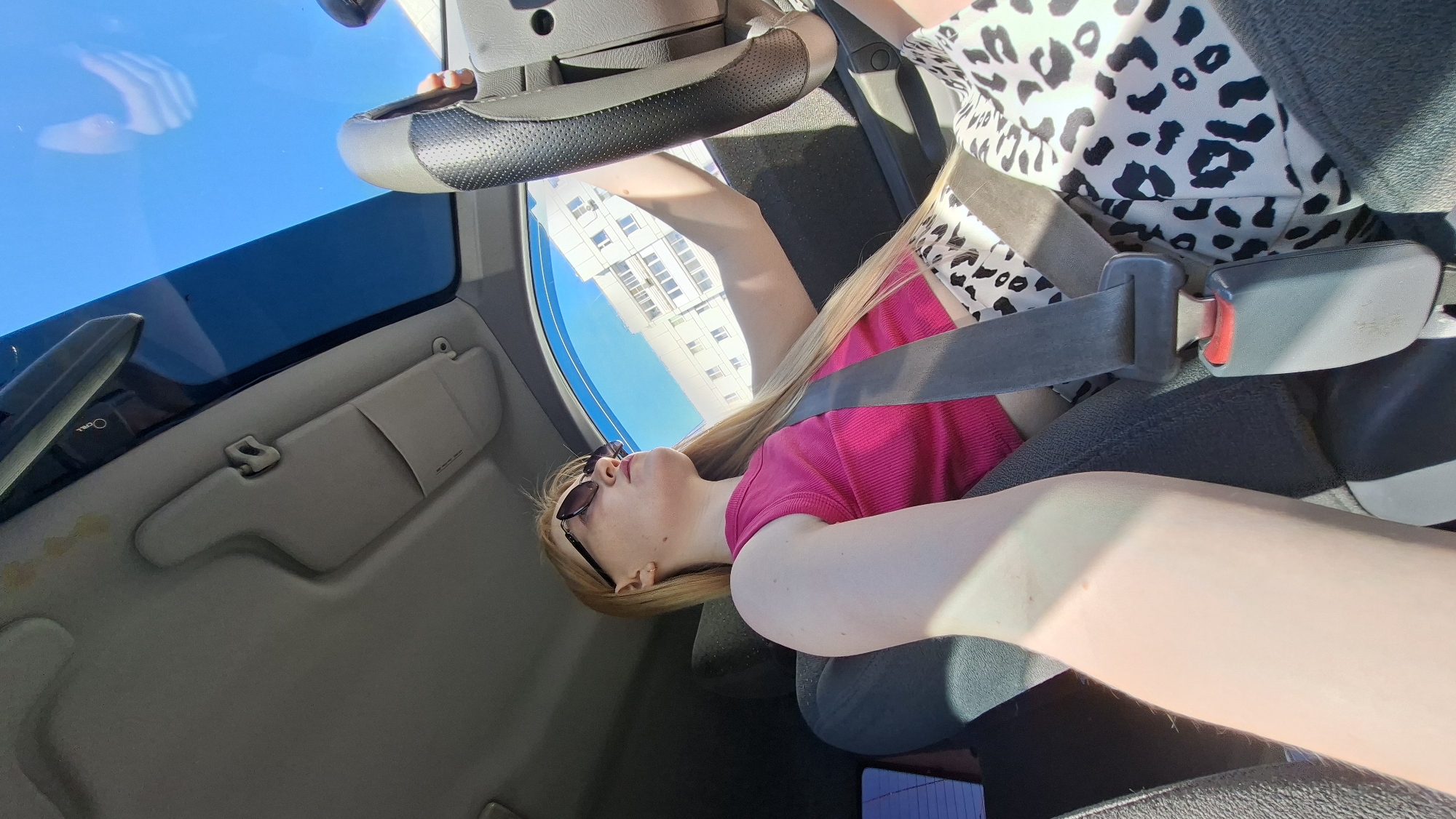 The image depicts a woman wearing sunglasses and a pink shirt, secured by a seatbelt in the passenger seat of a car. A white building is visible through the window, with the blue sky above. The location is Chaikovsky, Russia.