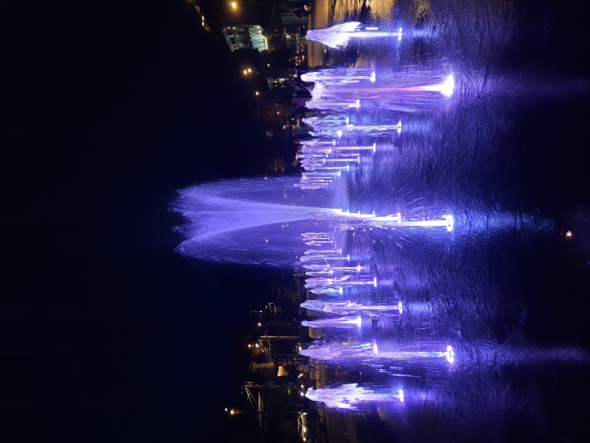 A night view of an illuminated fountain display in the city of Lviv, Ukraine. The fountain features numerous jets of water, each lit from below with purple and blue lights, creating a shimmering effect on the wet pavement. In the background, streetlights and building lights are visible, along with blurred figures of people walking and cars on a street. The sky is dark, indicating it is nighttime. The overall scene conveys a vibrant and dynamic urban nighttime atmosphere.