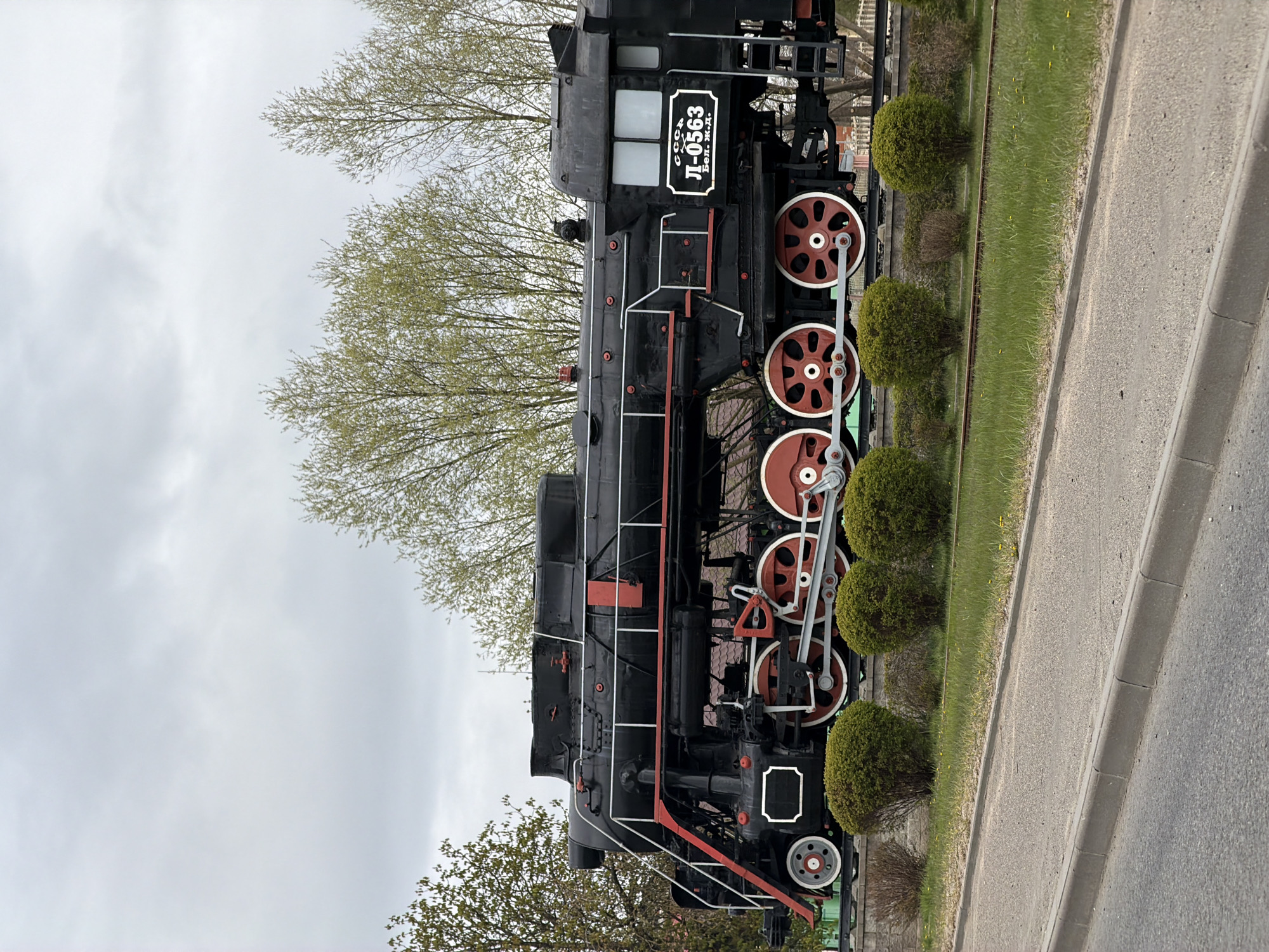 The image displays a large, black steam locomotive positioned as a static exhibit outdoors under an overcast sky. The train, an "L" series locomotive, features prominent red accents on its large driving wheels and connecting rods, as well as on various handrails and parts of its undercarriage. A white sign with black lettering is visible on the cab, reading "СССР" (USSR), "Л-0563" (L-0563), and "Бел. ж.д." (Bel. zh.d. – Belarusian Railway), indicating its origin and former service.
The locomotive is situated on what appears to be a section of railway track, flanked by a strip of green grass and several neatly trimmed, round green bushes. Beyond the grass and bushes, a paved road or pathway with a curb runs parallel to the train. In the background, numerous trees with light green, budding foliage suggest it is springtime. The sky is largely white and covered with clouds, indicating an overcast daytime. The overall scene suggests a quiet, public display in a park or similar open area in Lida, Belarus. The original image is rotated 90 degrees clockwise, making the ground appear on the right side and the sky on the left.