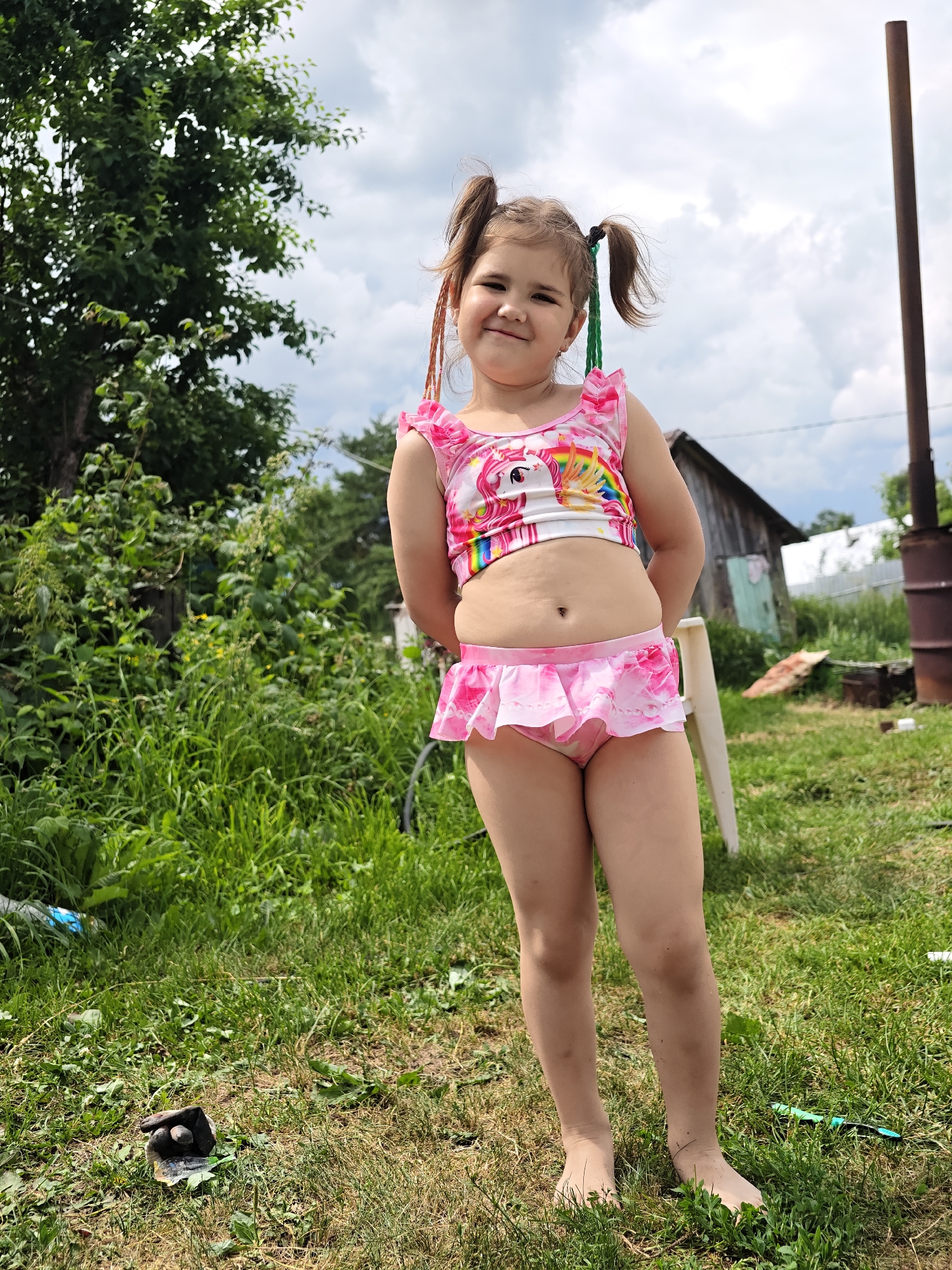 A young girl with pigtails is standing in a grassy outdoor area in Noviy, Russia. She is wearing a pink and white swimsuit with a unicorn and rainbow design. The swimsuit includes a ruffled top and a skirted bottom. She has her hands behind her back and is smiling at the camera. The background shows a wooden structure, some trees, and a cloudy sky. The time of day appears to be daytime. There is no visible text in the image.