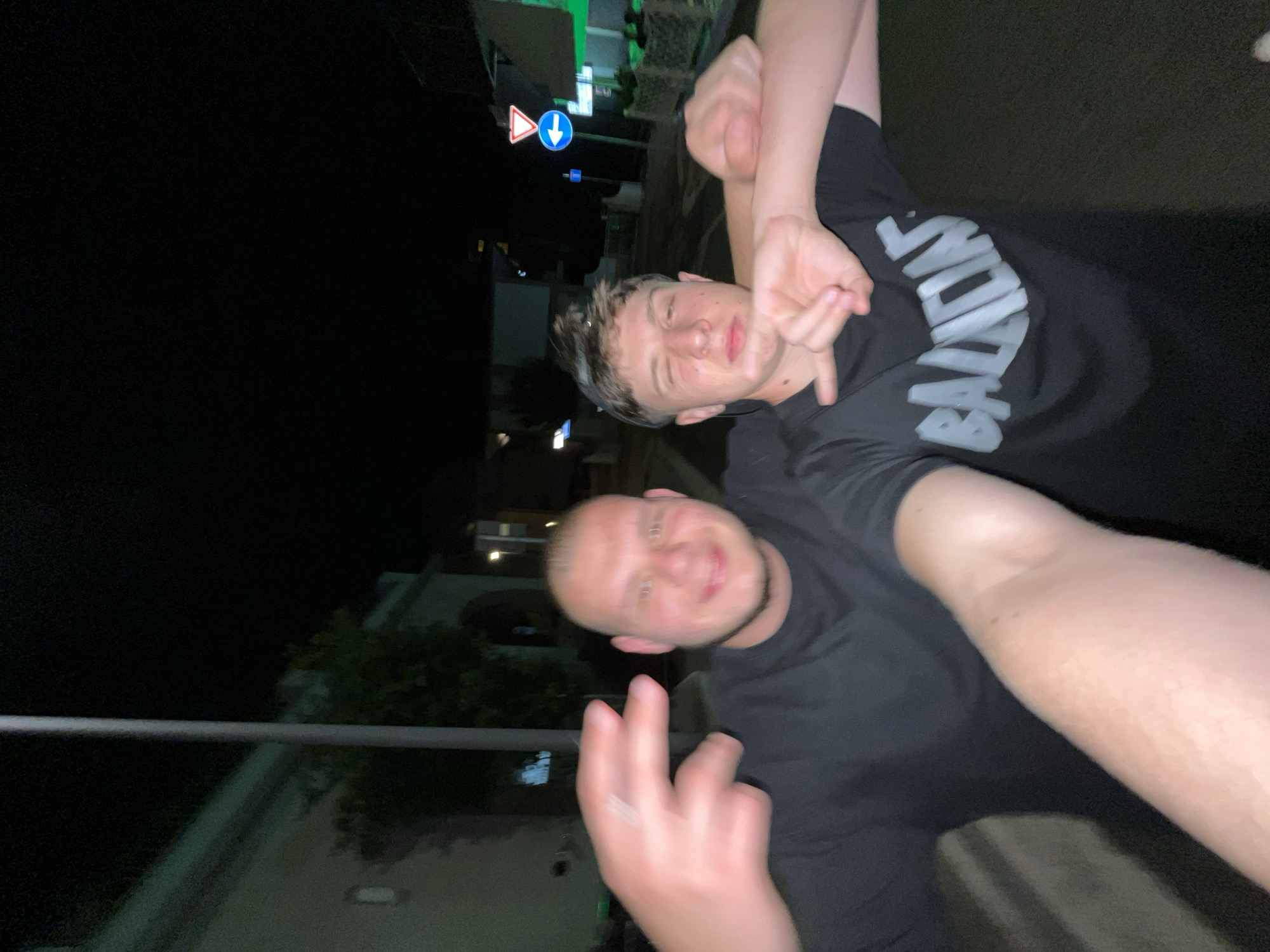 Two people pose for a selfie at night on a street in Jesolo, Italy. The person in the foreground holds the camera at arm's length, showing two fingers. The second person is behind, their arm around the first person's neck. Both people wear dark t-shirts. The word "BALMAIN" is visible on the second person's shirt. A traffic sign is visible in the background.