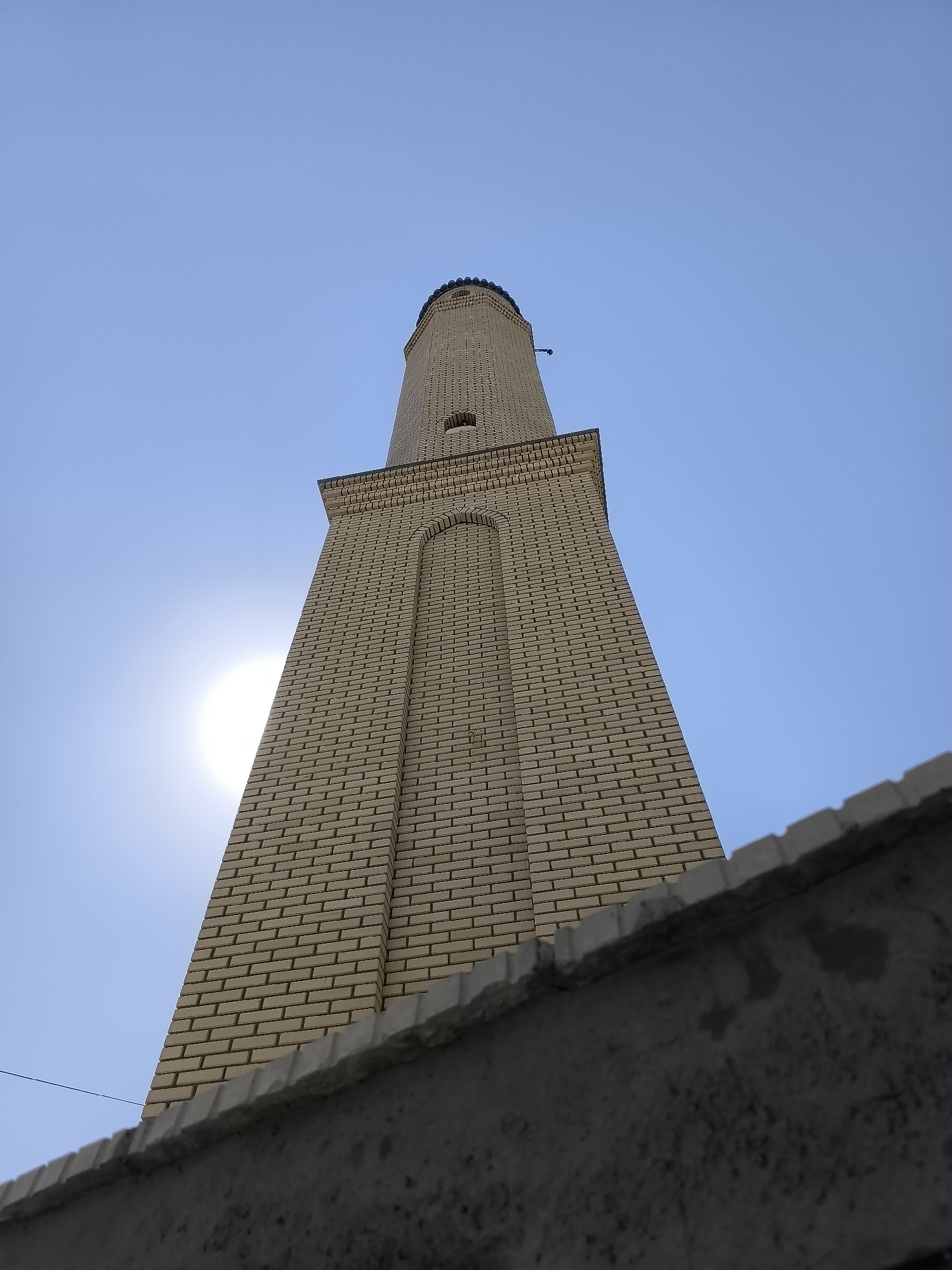 An upward-angle shot shows a tall, narrow minaret made of light-colored bricks against a clear blue sky. A portion of the sun is visible behind the minaret, casting a glare. The minaret has several levels, with distinct architectural details such as arched windows and decorative brickwork near the top. At the very top, a dark, rounded cap is visible. In the foreground, the top of a rough, gray concrete wall runs horizontally across the bottom of the frame, partially obscuring the base of the minaret and creating a sense of depth. A thin black wire is visible stretching across the scene from the left side.