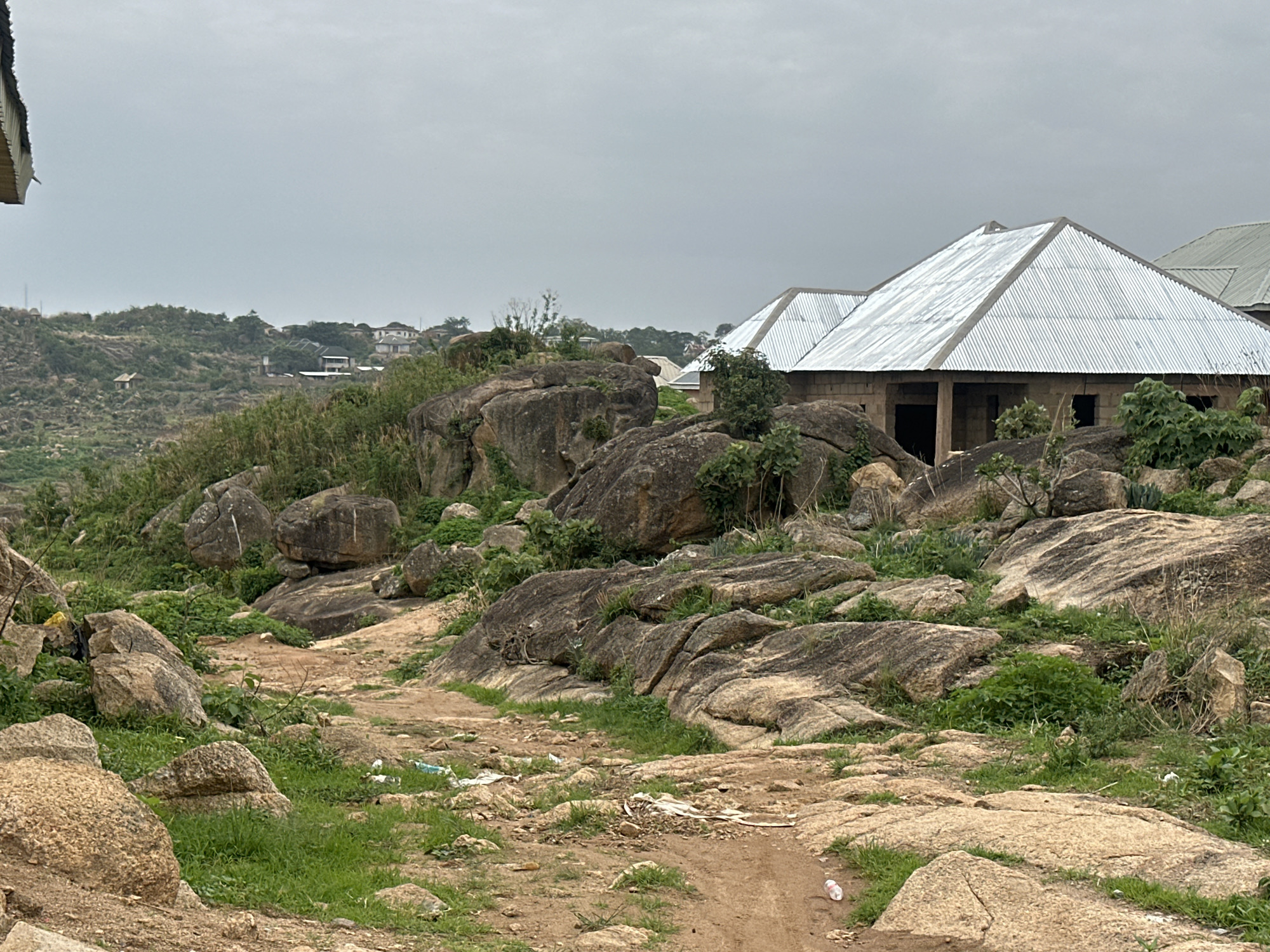 The image depicts a rocky, uneven terrain leading towards unfinished buildings in Jos, Nigeria. Large boulders interspersed with sparse vegetation dominate the landscape. A dirt path winds through the rocks, littered with debris. In the background, more buildings are visible on a slight incline. The sky is overcast.